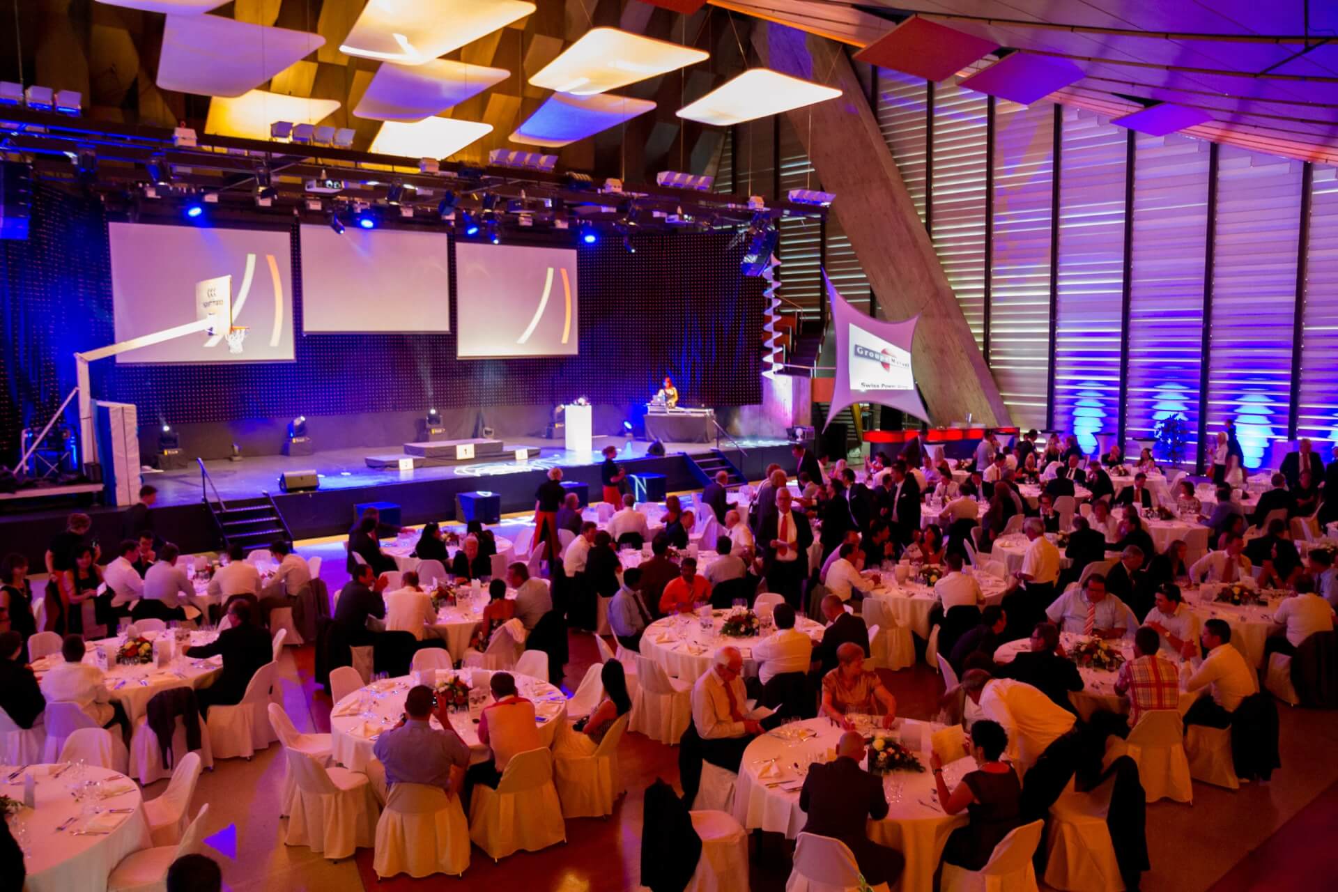 Eine große formelle Veranstaltung mit runden Tischen, sitzenden Gästen und einer Bühne mit Bildschirmen an der Vorderseite, bei violetter Beleuchtung in einem modernen Veranstaltungsort.