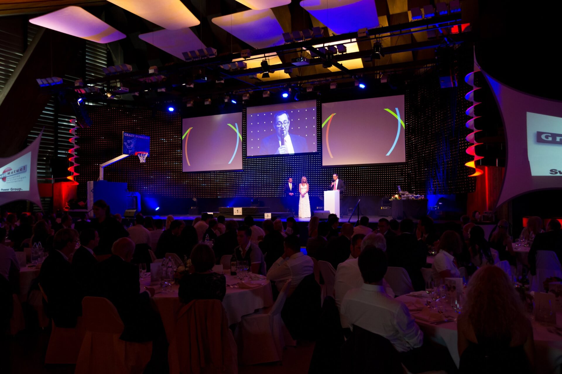 Eine formelle Veranstaltung mit sitzenden Gästen, die einer Bühnenpräsentation beiwohnen; zwei Personen stehen auf der Bühne, hinter ihnen wird ihr Bild auf einer großen Leinwand gezeigt.