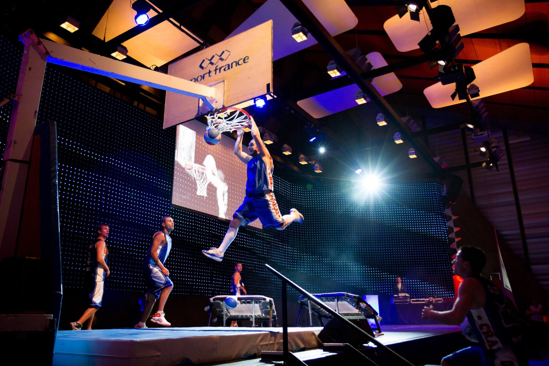 Ein Basketballspieler springt von einem Trampolin und wirft den Ball während eines Spiels in einer Halle, während andere Spieler zusehen und das Spielfeld hell erleuchtet ist.