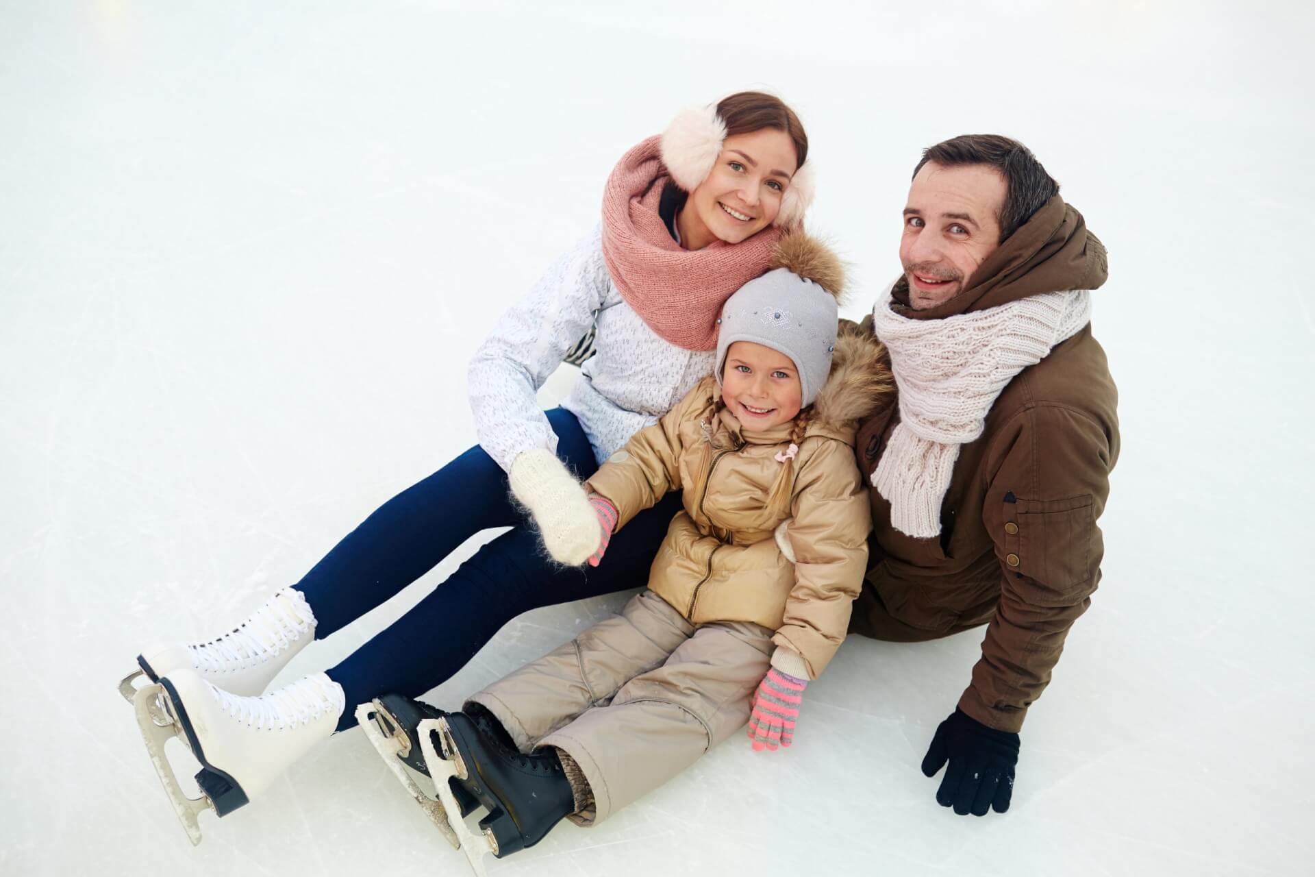 Drei Personen in Winterkleidung und Schlittschuhen sitzen auf einer Eisbahn und lächeln in die Kamera.