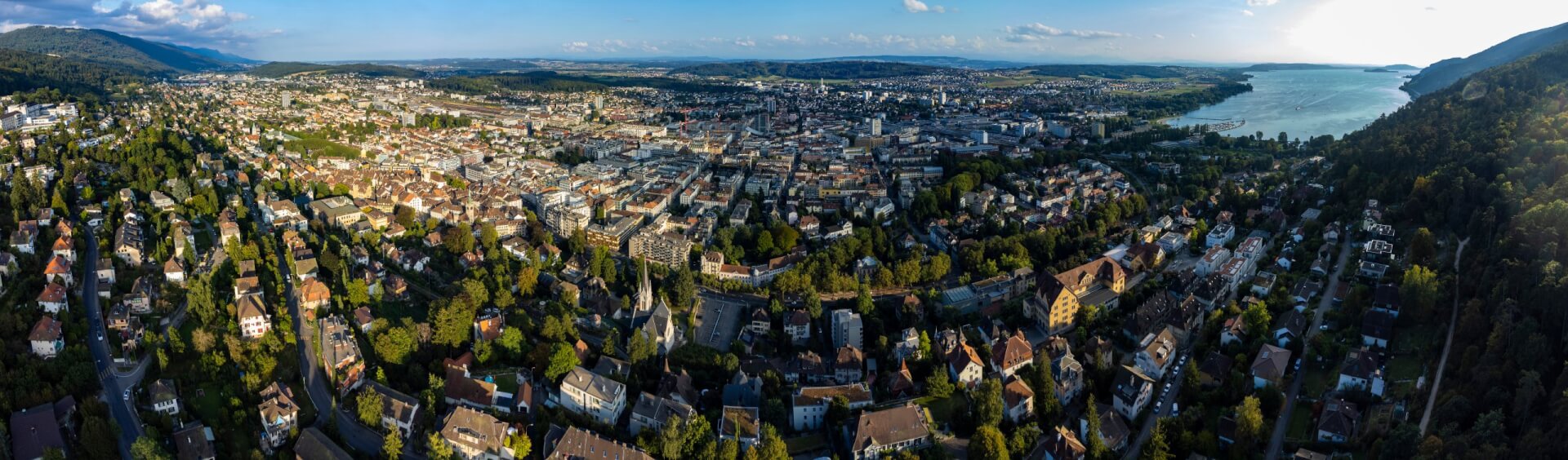 Panoramablick aus der Luft auf eine Stadt mit dichter Bebauung, Grünflächen und einem großen, von Hügeln gesäumten See unter einem teilweise bewölkten Himmel.
