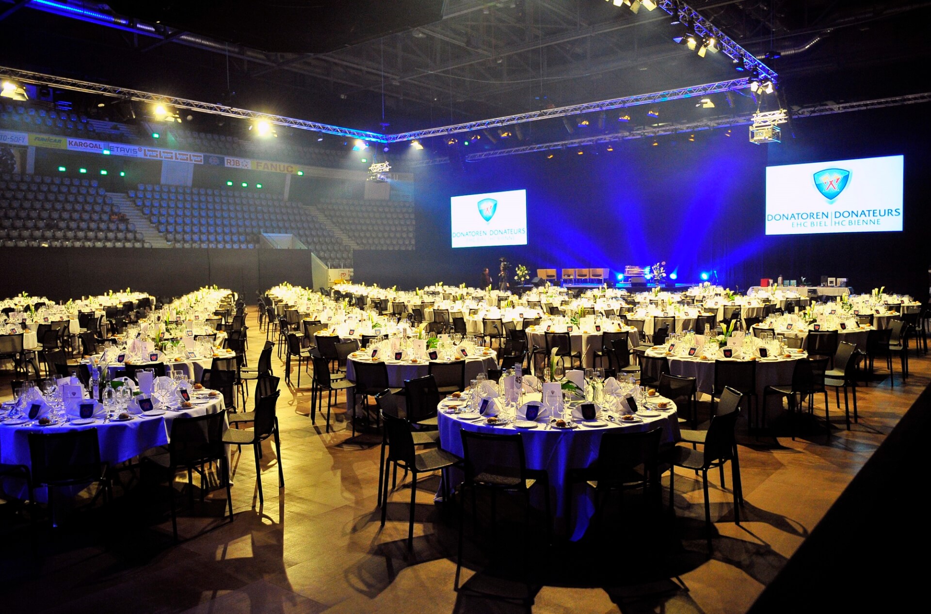 Ein großer Bankettsaal mit runden Tischen, die für eine Veranstaltung gedeckt sind, eine Bühne mit blauer Beleuchtung und Projektionsflächen, auf denen Donatoren Donateurs zu sehen sind.
