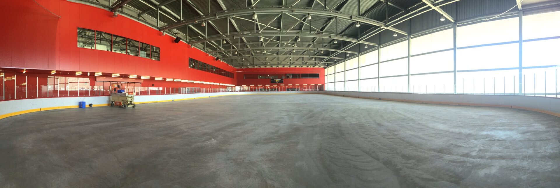 Weitwinkelansicht einer leeren Eissporthalle mit roter Wand, großen Fenstern und freiliegenden Deckenbalken.