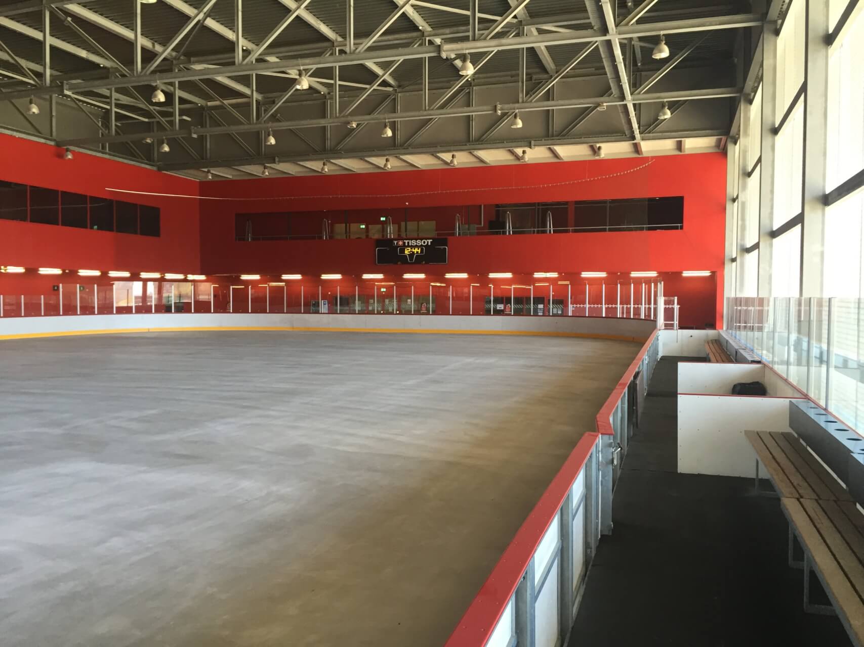 Eine Eishalle ohne Eis, umgeben von roten Wänden und Glasbarrieren, mit leeren Bänken und einer Anzeigetafel im Hintergrund.