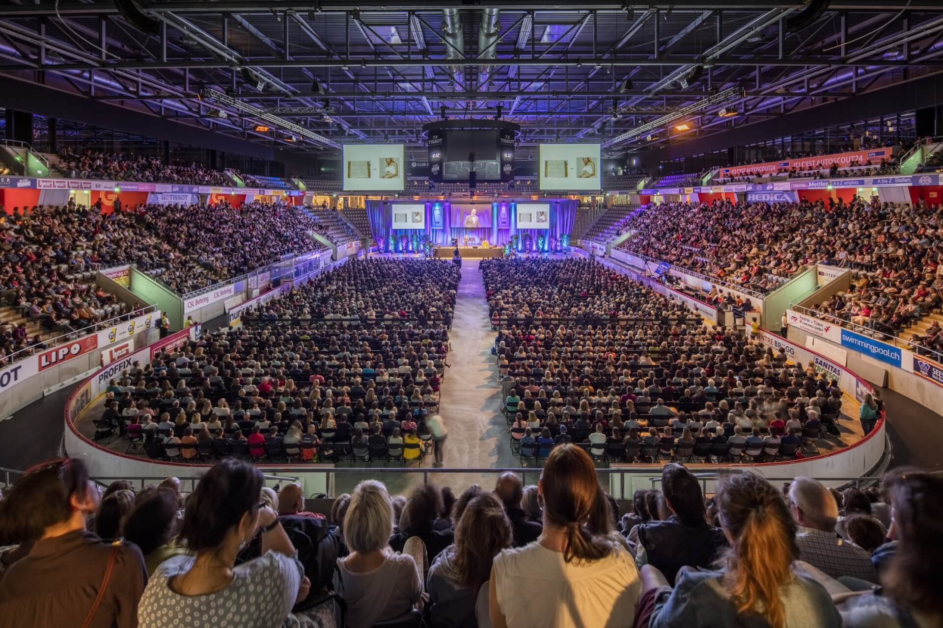Ein großes Publikum füllt eine überdachte Arena, alle blicken auf eine hell erleuchtete Bühne am anderen Ende, mit Sitzreihen auf beiden Seiten und Bannern an den Wänden.