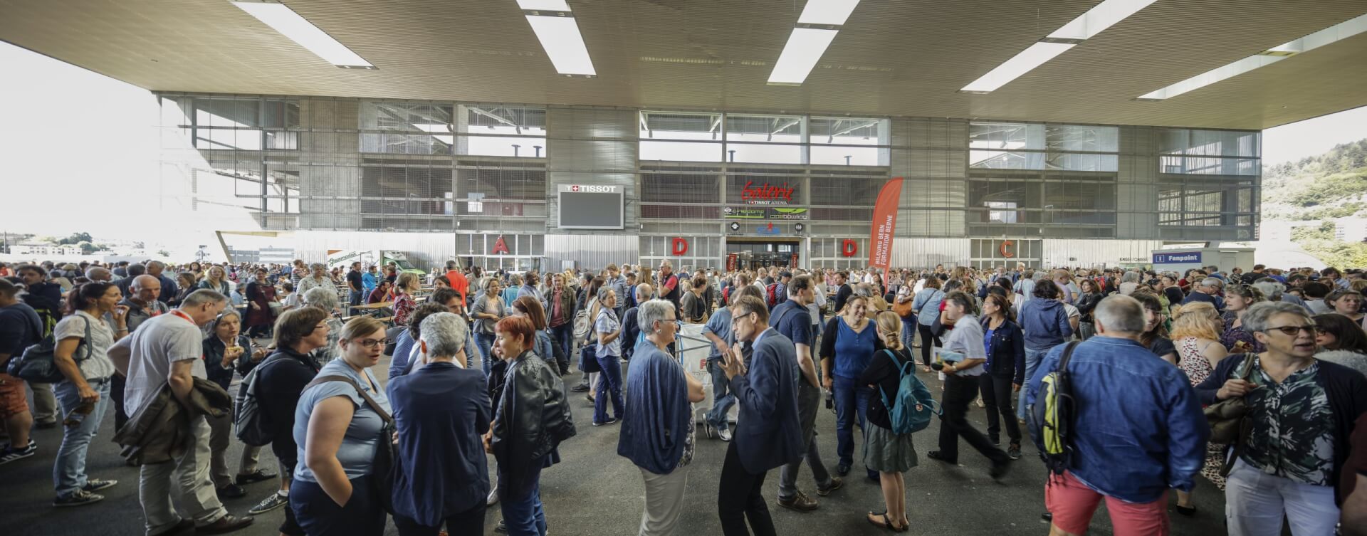 Eine große Menschenmenge steht vor einem modernen Gebäude unter einem überdachten Bereich, unterhält sich und wartet.