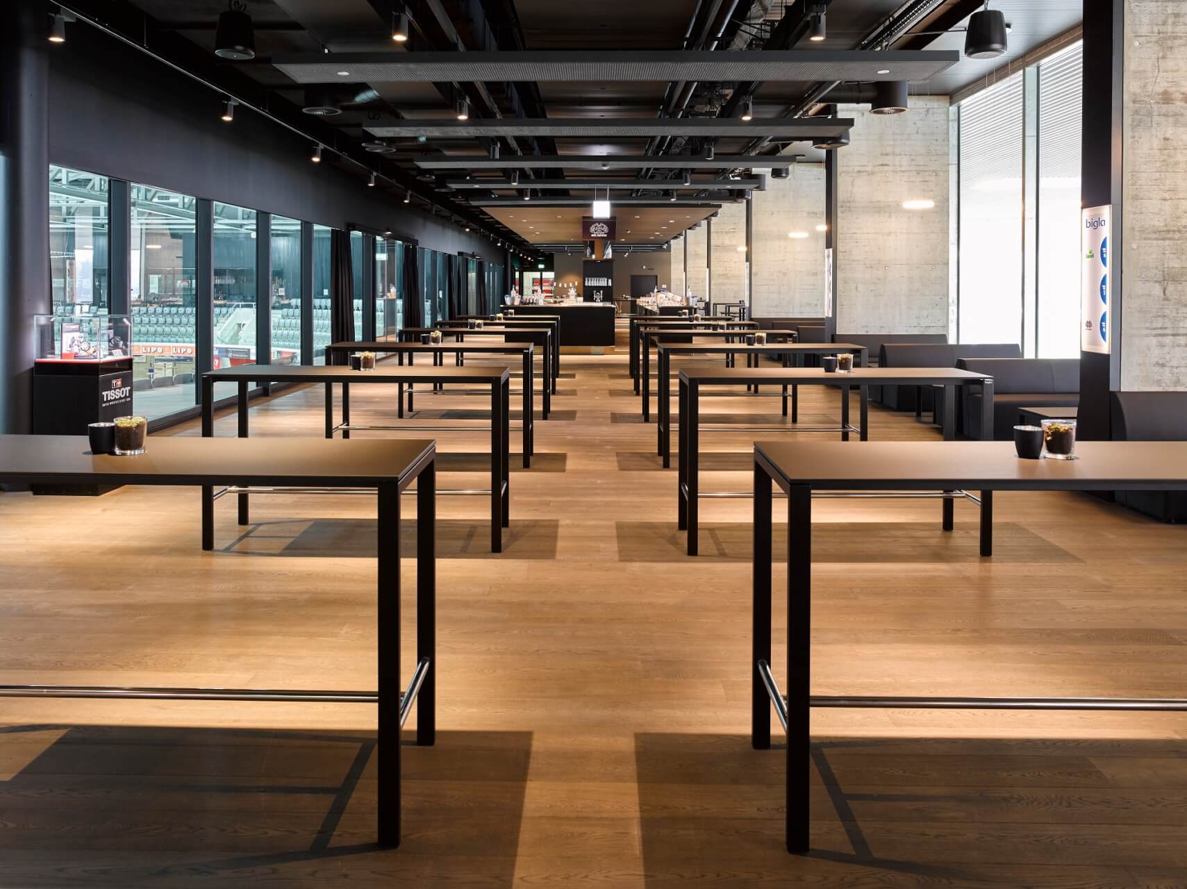 Ein moderner, leerer Essbereich mit schwarzen Tischen und Stühlen, die in Reihen auf einem Holzboden angeordnet sind, mit großen Fenstern und Beleuchtung im industriellen Stil.