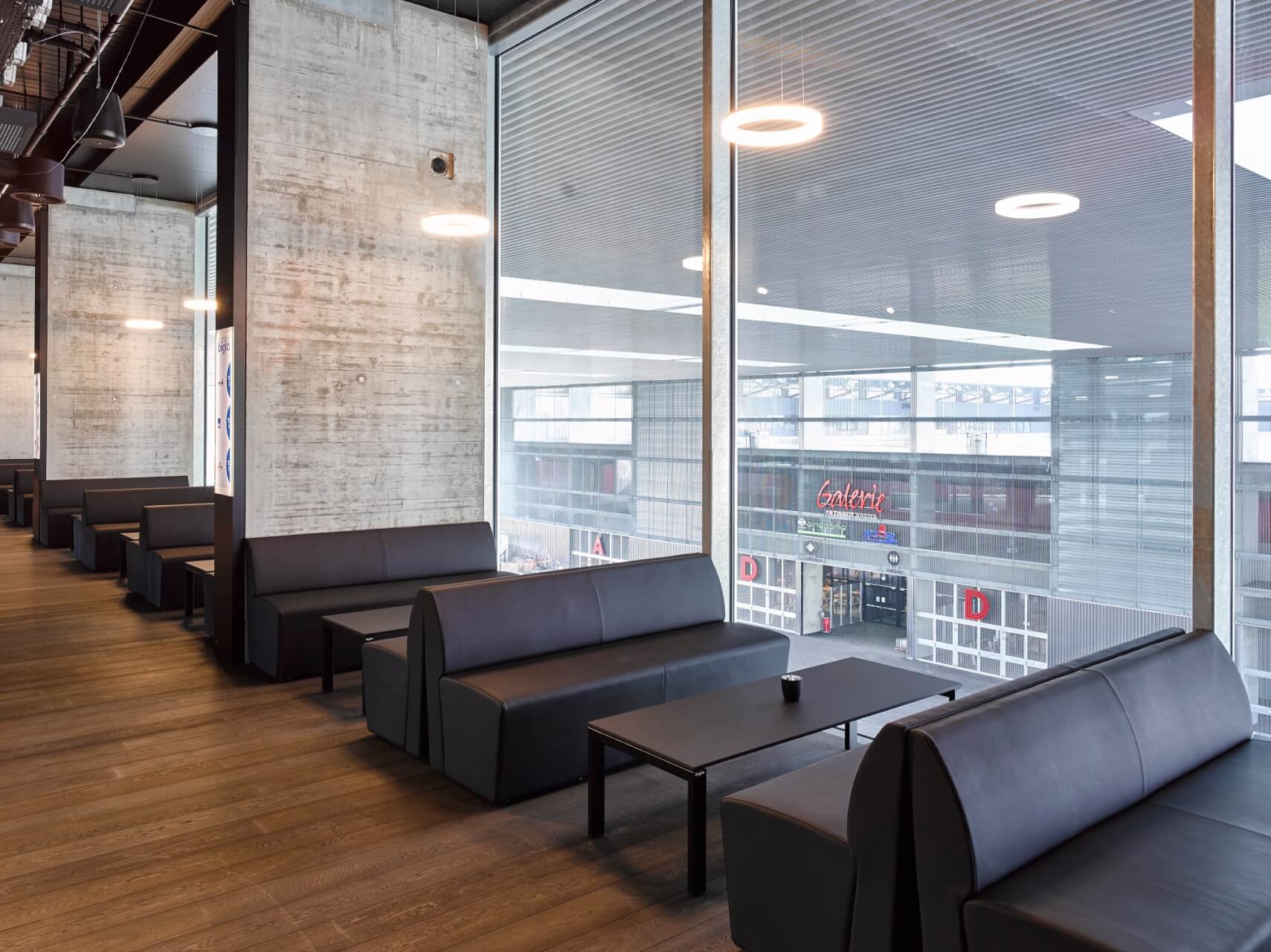 Moderner Lounge-Bereich mit schwarzen Sofas und Tischen neben großen Fenstern, mit Blick auf ein Einkaufszentrum mit sichtbarer Beschilderung.