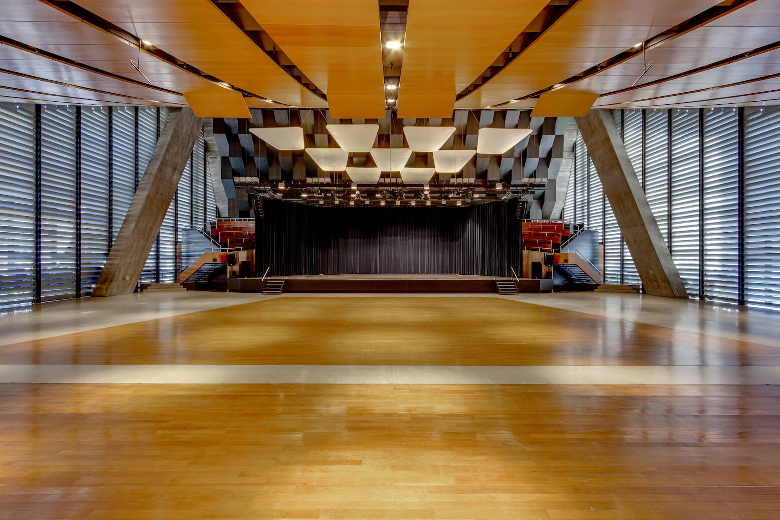 Ein geräumiges, modernes Auditorium mit Holzboden, seitlichen Sitzreihen, schrägen Wänden und einer Bühne mit dunklen Vorhängen im hinteren Teil.