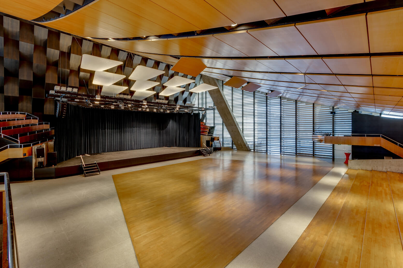 Modernes Auditorium mit Holzfußboden, geometrischen Deckenpaneelen, einer großen Bühne mit schwarzen Vorhängen und abgestuften Sitzreihen an einer Seite. Durch die raumhohen Fenster fällt natürliches Licht ein.