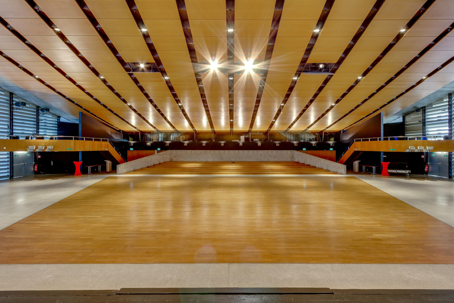 Geräumige, leere, moderne Halle mit Holzboden, Sitzreihen im hinteren Bereich und einer Decke mit Lichtbändern.