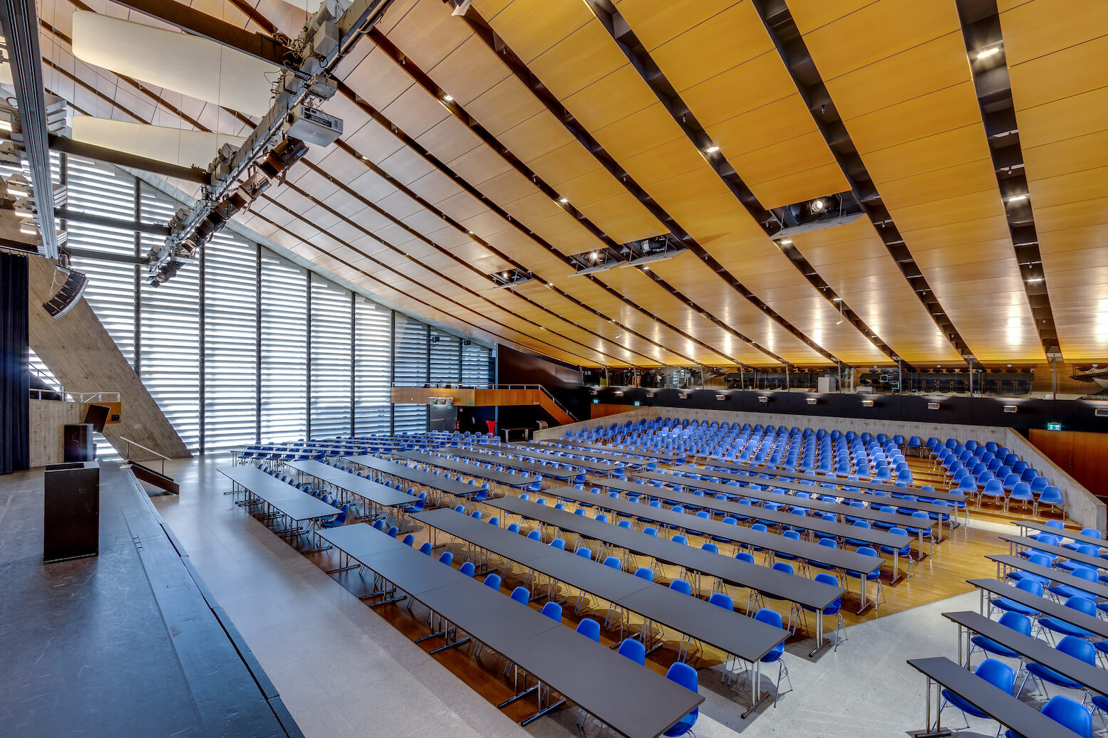Großer moderner Hörsaal mit blauen Stuhlreihen und langen Tischen, holzgetäfelter Decke und einer breiten Bühne auf der linken Seite. Natürliches Licht strömt durch hohe Fenster.