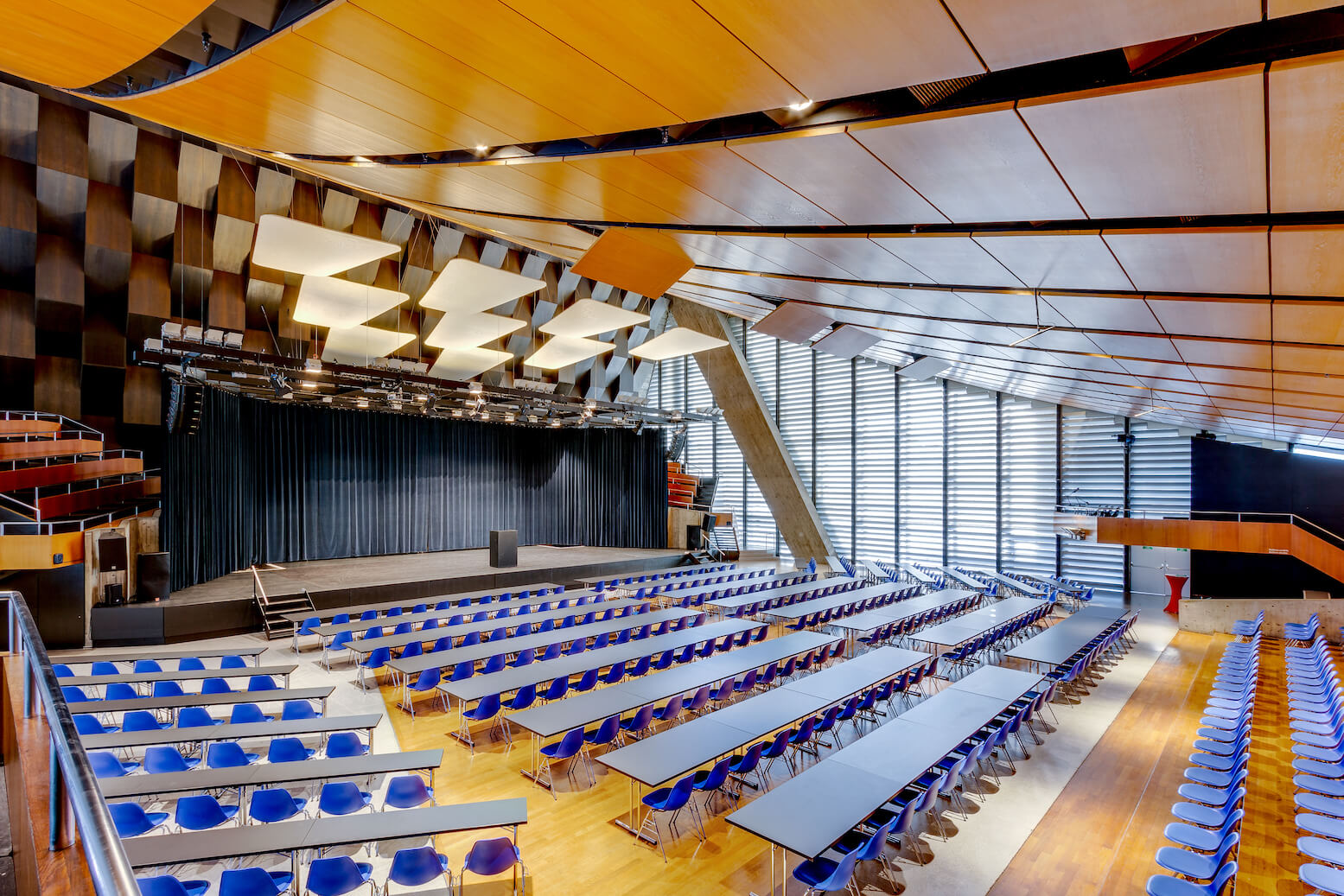 Großes, modernes Auditorium mit blauen Stuhlreihen und langen Tischen vor einer Bühne, mit holzgetäfelter Decke und großen Fenstern auf einer Seite.