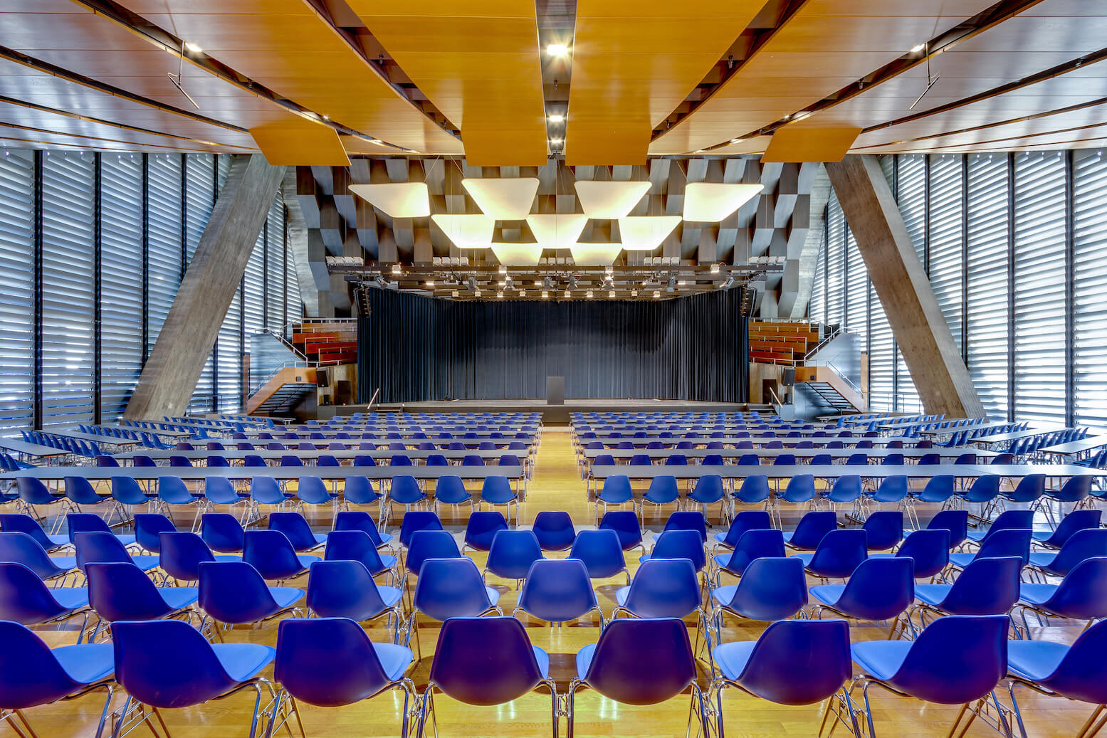 Ein großes Auditorium mit blauen Stuhlreihen vor einer Bühne mit Podium, unter geometrischen Deckenplatten und flankiert von hohen Fenstern.