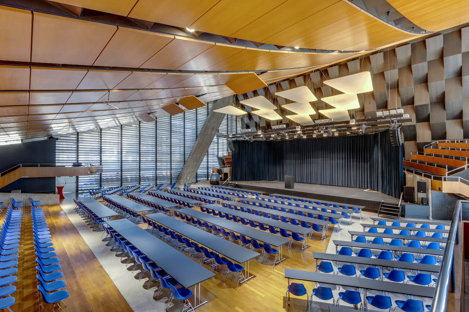 Modernes Auditorium mit langen Reihen blauer Stühle und Tische vor einer Bühne mit schwarzen Vorhängen, mit Holzfußboden und geometrischen Deckenplatten.