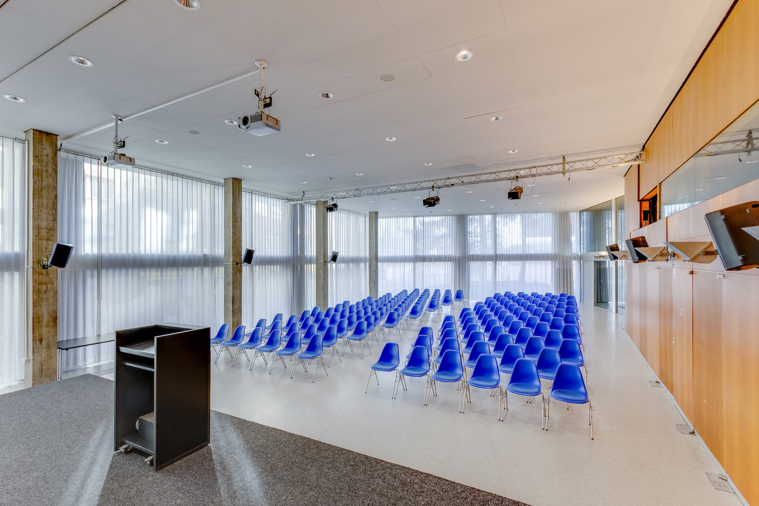 Ein moderner Konferenzraum mit blauen Stuhlreihen vor einem Podium und einer Projektionsfläche, umgeben von raumhohen Fenstern und Holzverkleidungen.