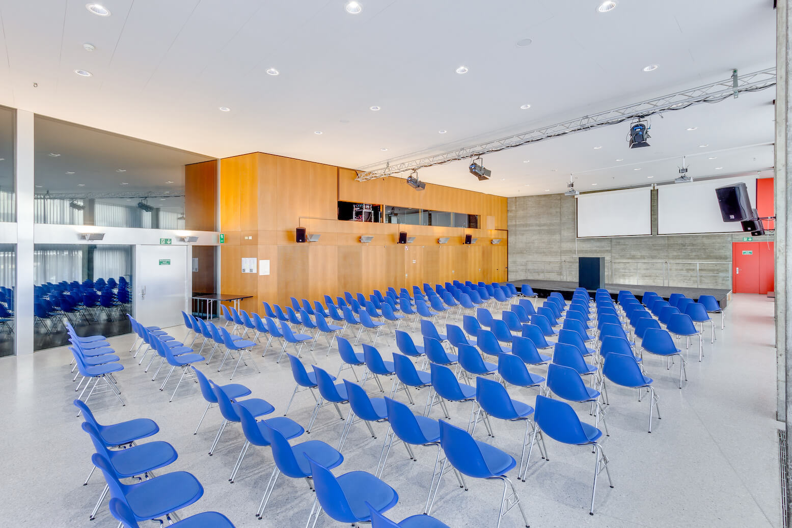In einem modernen, hellen Konferenzraum mit Holzvertäfelung, Betonwänden und an der Decke installierten audiovisuellen Geräten sind Reihen leerer blauer Stühle aufgestellt.