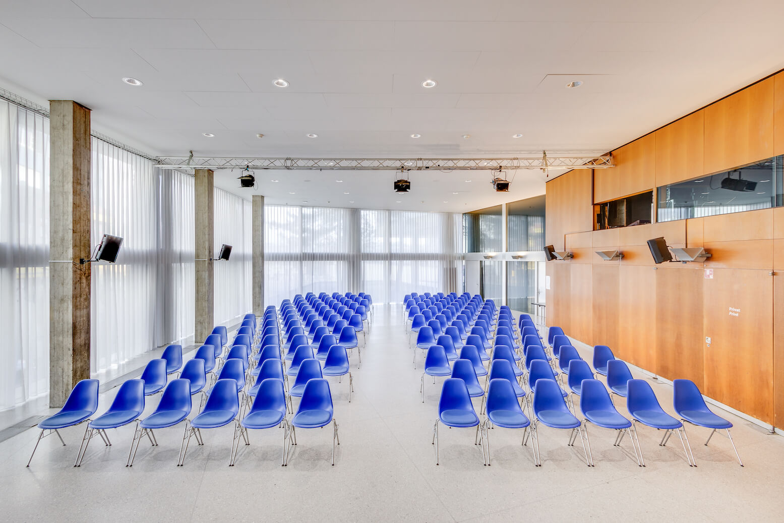 In einem modernen, hellen Konferenzraum mit großen Fenstern und Holzvertäfelung stehen leere blaue Stühle in Reihen nach vorne gerichtet.