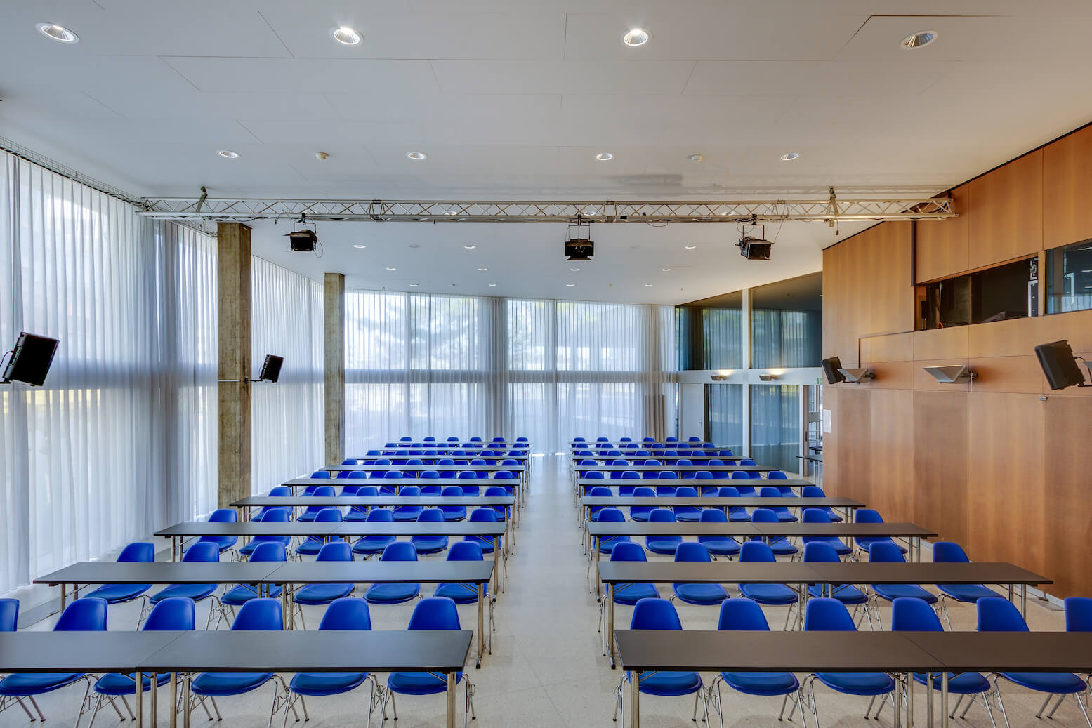 Modernes Klassenzimmer mit blauen Stuhlreihen und schwarzen Tischen an der Vorderseite, großen Fenstern mit transparenten Vorhängen und hölzernen Wandpaneelen auf der rechten Seite.