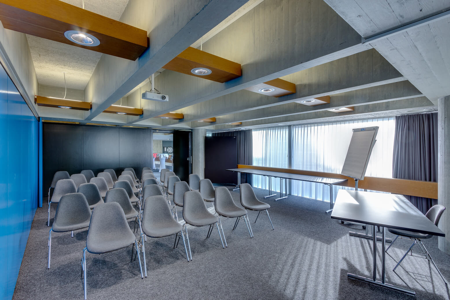 Moderner Konferenzraum mit grauen Stuhlreihen vor einer Projektionsfläche, einem Flipchart, langen Tischen und großen Fenstern mit transparenten Vorhängen.