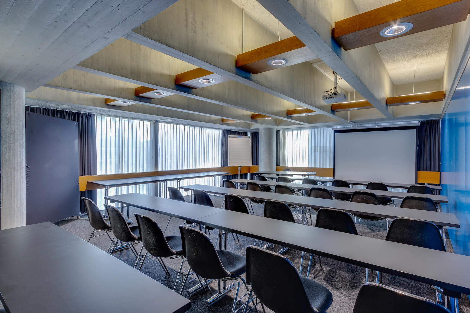 Ein moderner, leerer Konferenzraum mit Reihen schwarzer Stühle und langen Tischen, die vor einem Projektionsschirm und einem Flipchart stehen, umgeben von großen Fenstern mit Vorhängen.