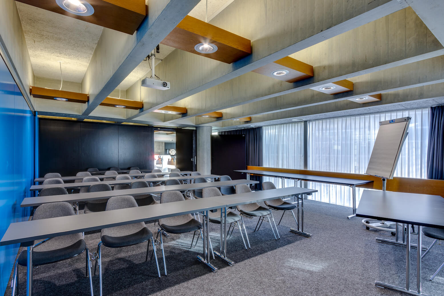 Ein moderner Konferenzraum mit Reihen von grauen Stühlen und Tischen, einem Flipchart, einem Projektor an der Decke und großen Fenstern mit durchsichtigen Vorhängen.