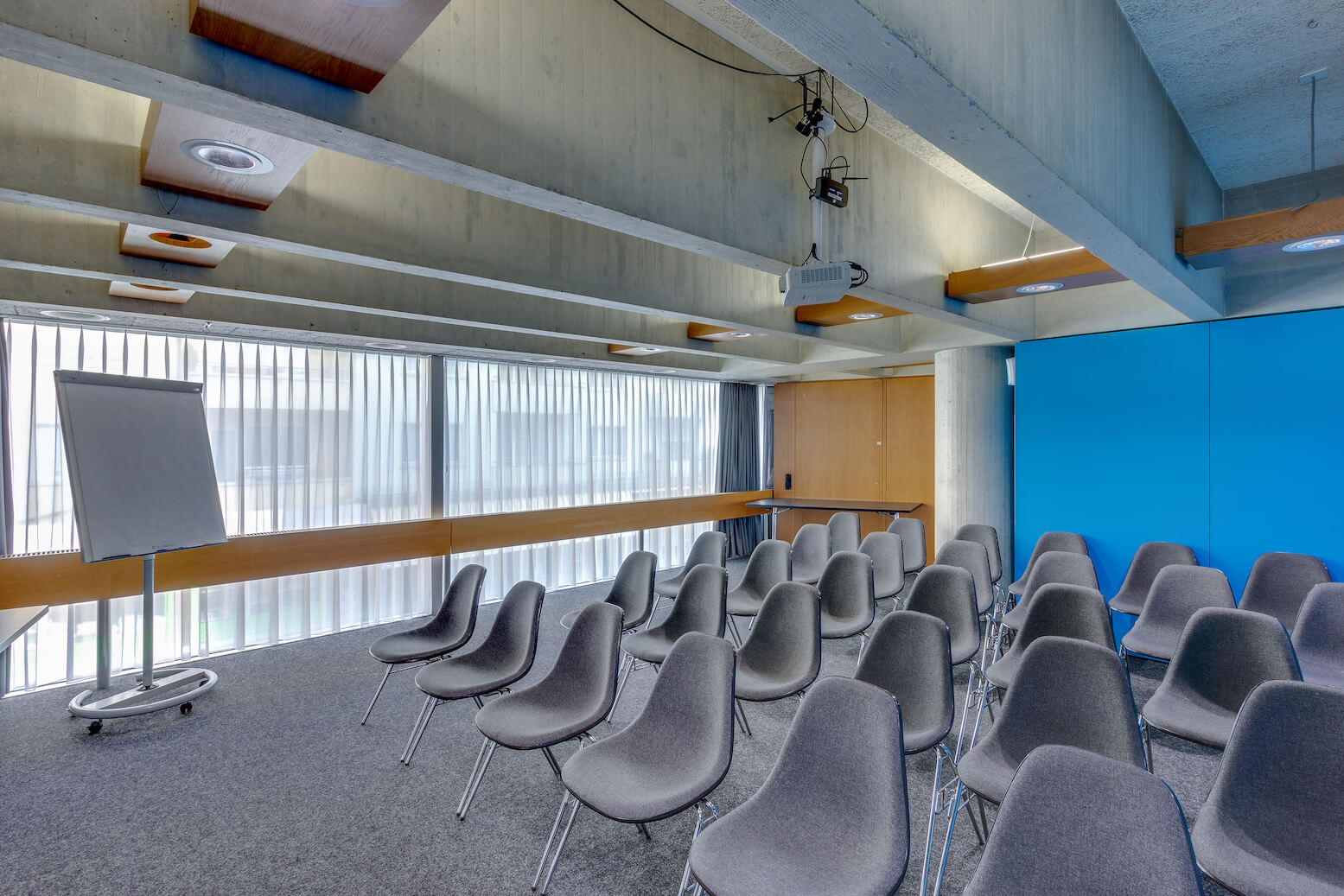 Ein kleiner Konferenzraum mit leeren grauen Stühlen, einem Flipchart, großen Fenstern mit Jalousien und einer blauen Akzentwand.