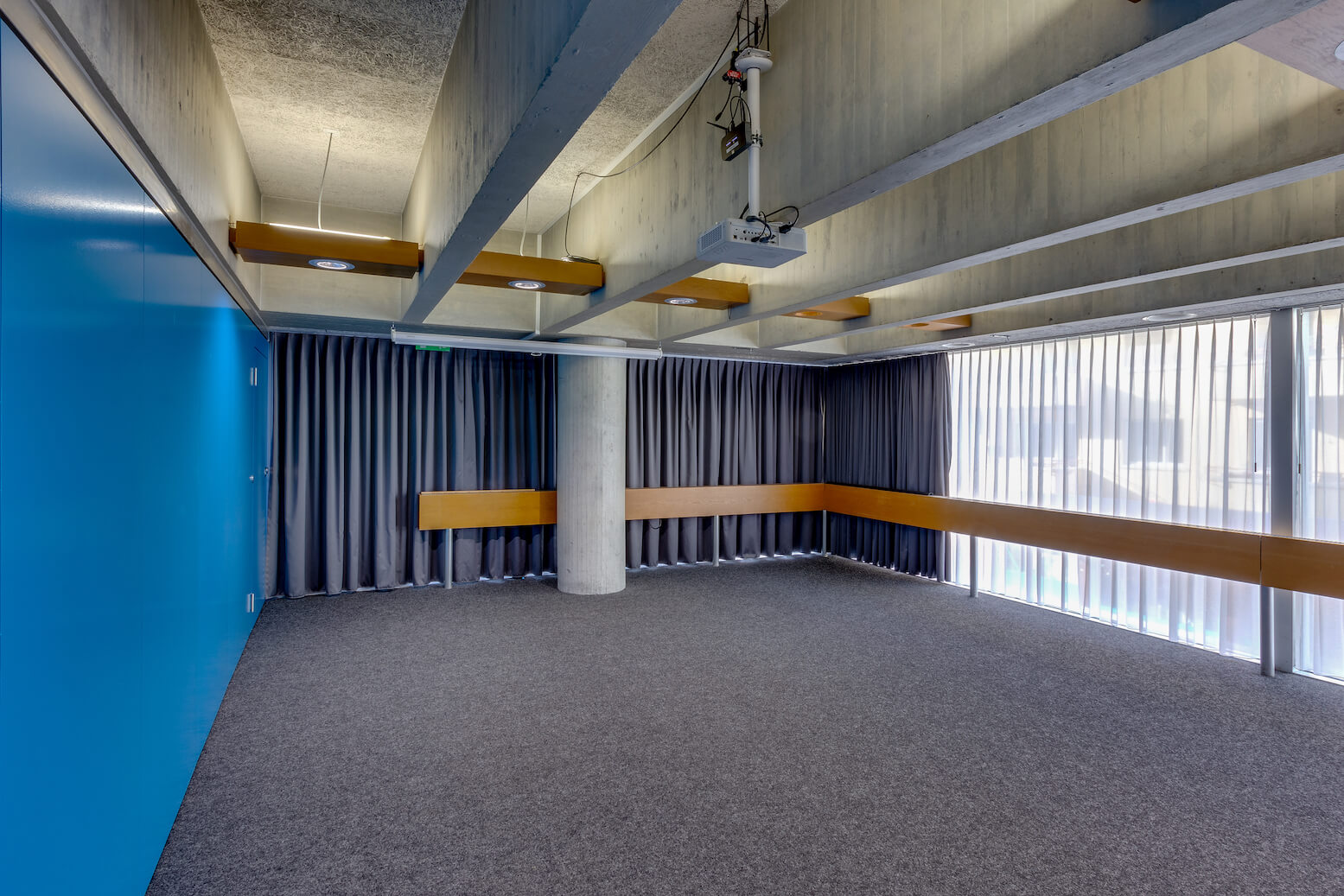 Leerer moderner Raum mit grauem Teppich, blauer Wand, Sichtbetondecke, Holzakzenten, Deckenprojektor, grauen Vorhängen und großen vertikalen Fenstern.