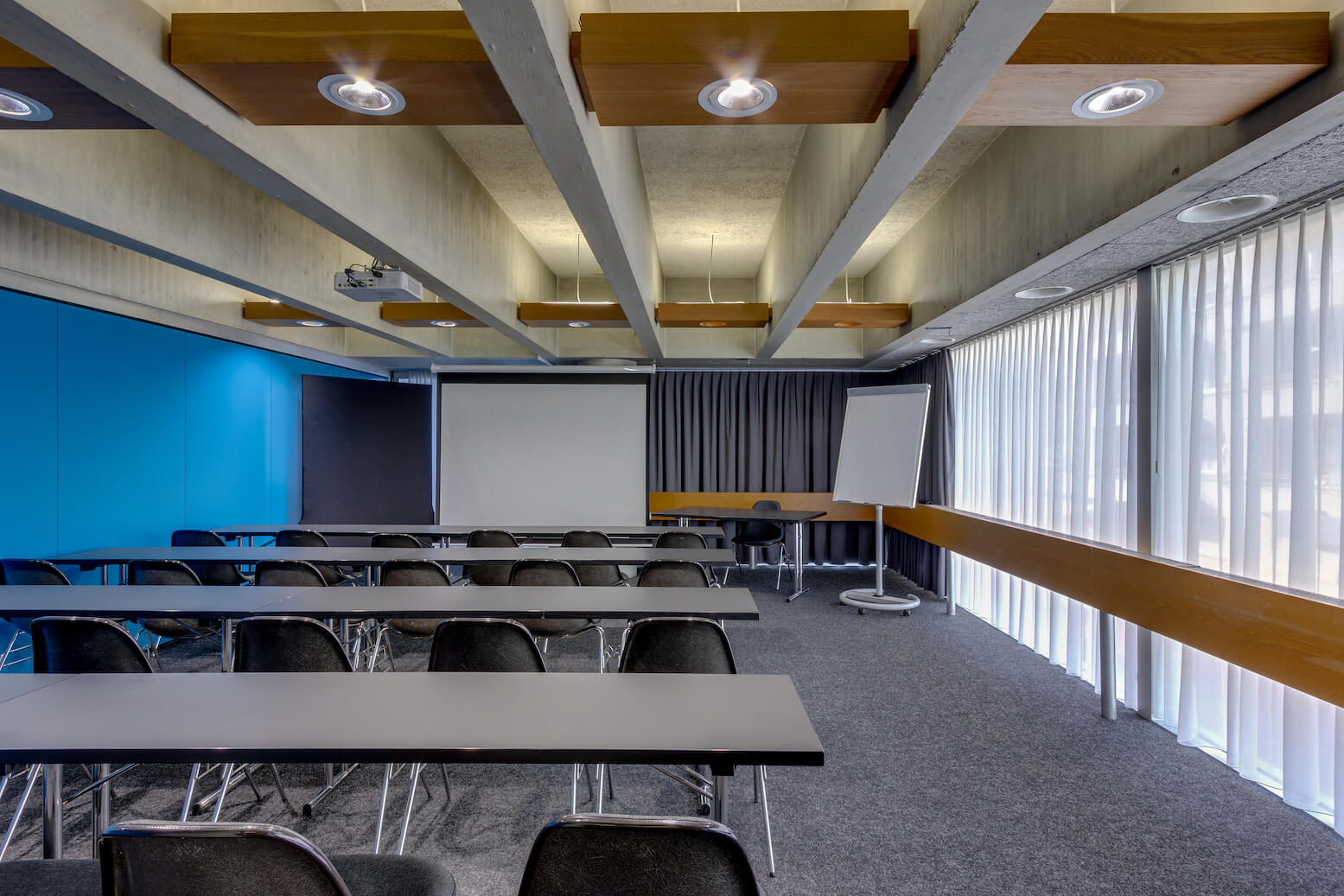 Modernes Klassenzimmer mit Reihen schwarzer Stühle und Tische vor einer Projektionsfläche, Flipchart und Fenstern mit vertikalen Jalousien. An der Decke befinden sich sichtbare Balken und Hängelampen.