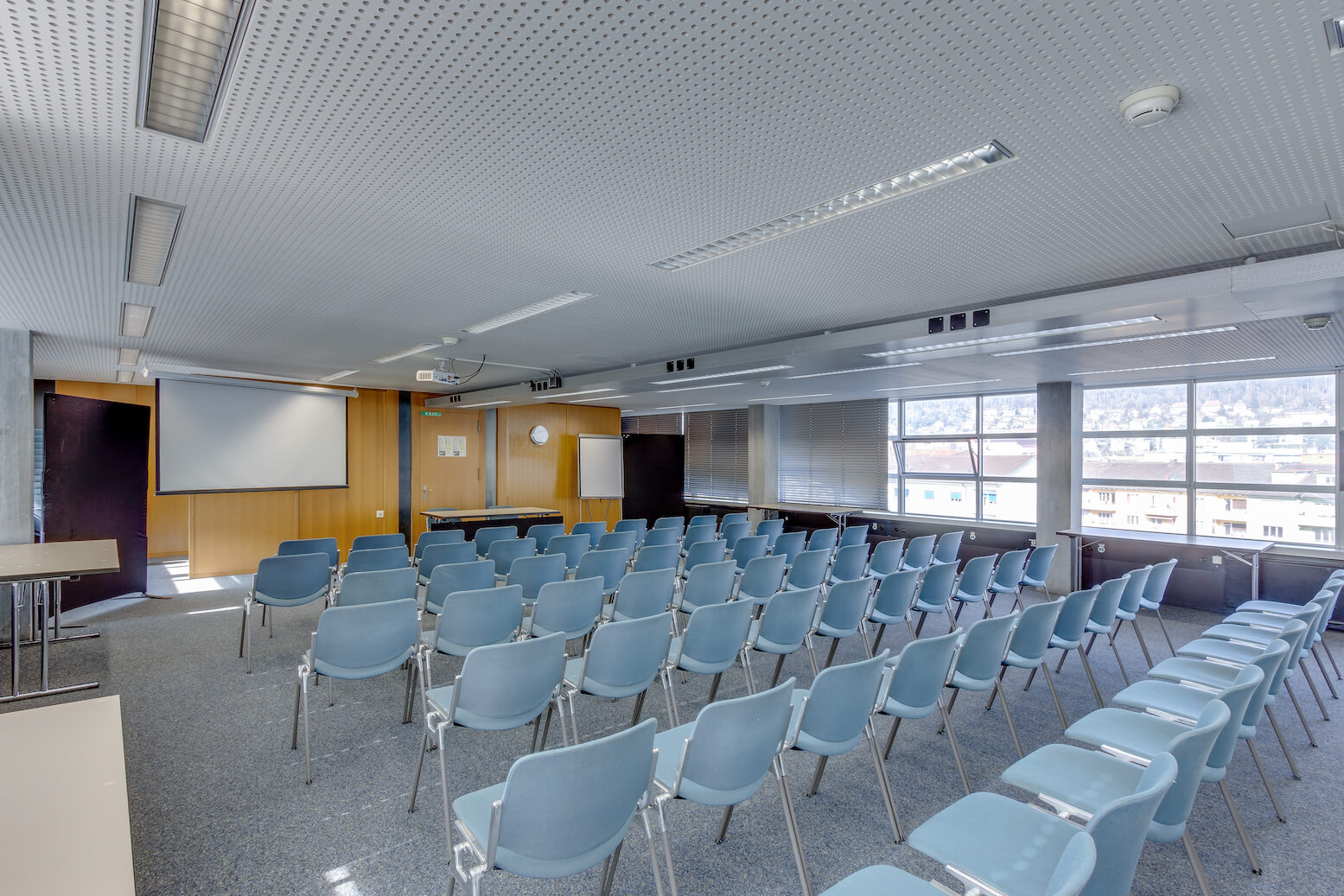 In einem modernen Konferenz- oder Vortragsraum mit großen Fenstern und Teppichboden stehen leere blaue Stühle vor einer Projektionsfläche.
