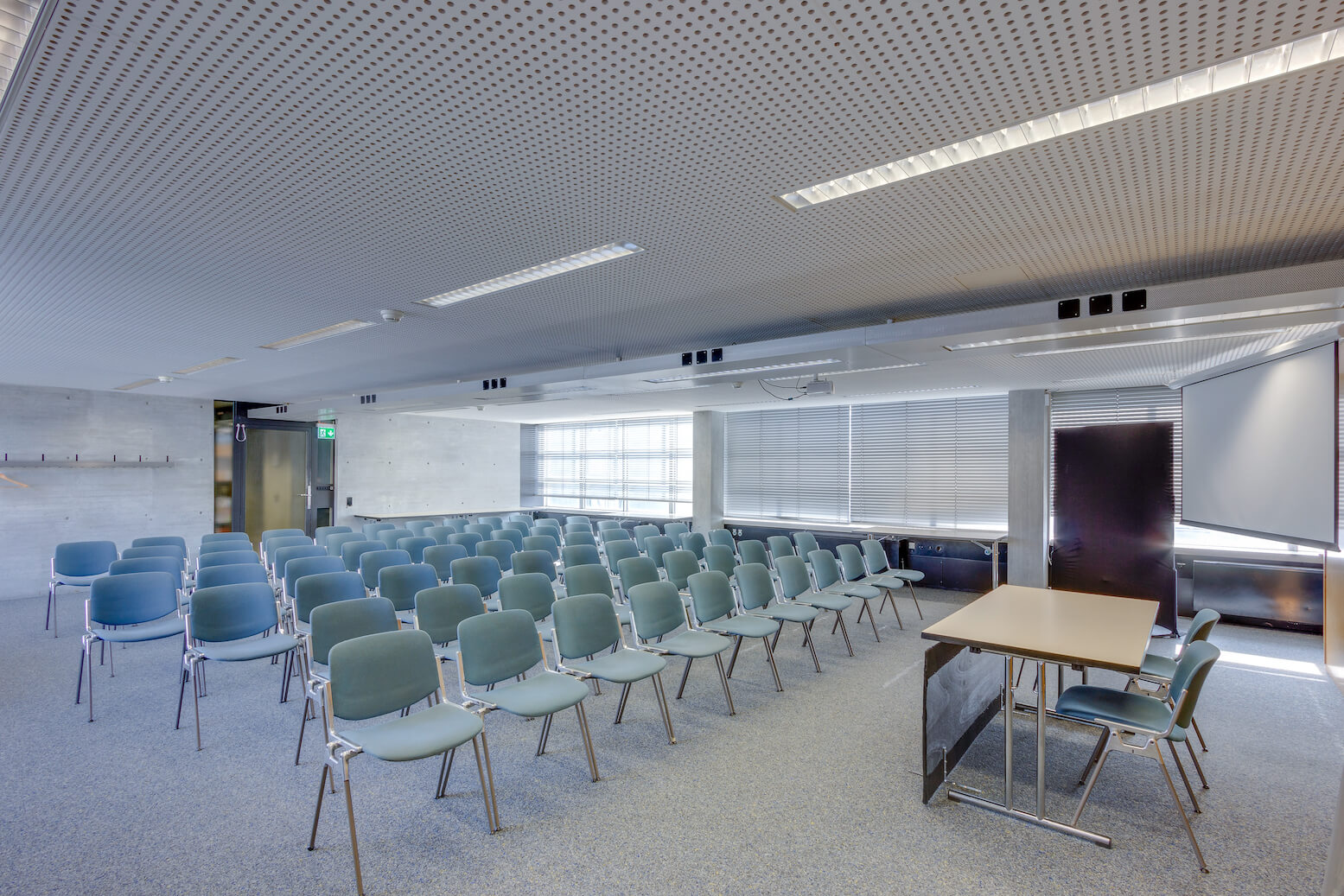 Ein moderner, leerer Konferenzraum mit Stuhlreihen vor einem Tisch und einer Projektionsfläche; große Fenster säumen eine Wand, und der Raum ist hell erleuchtet.