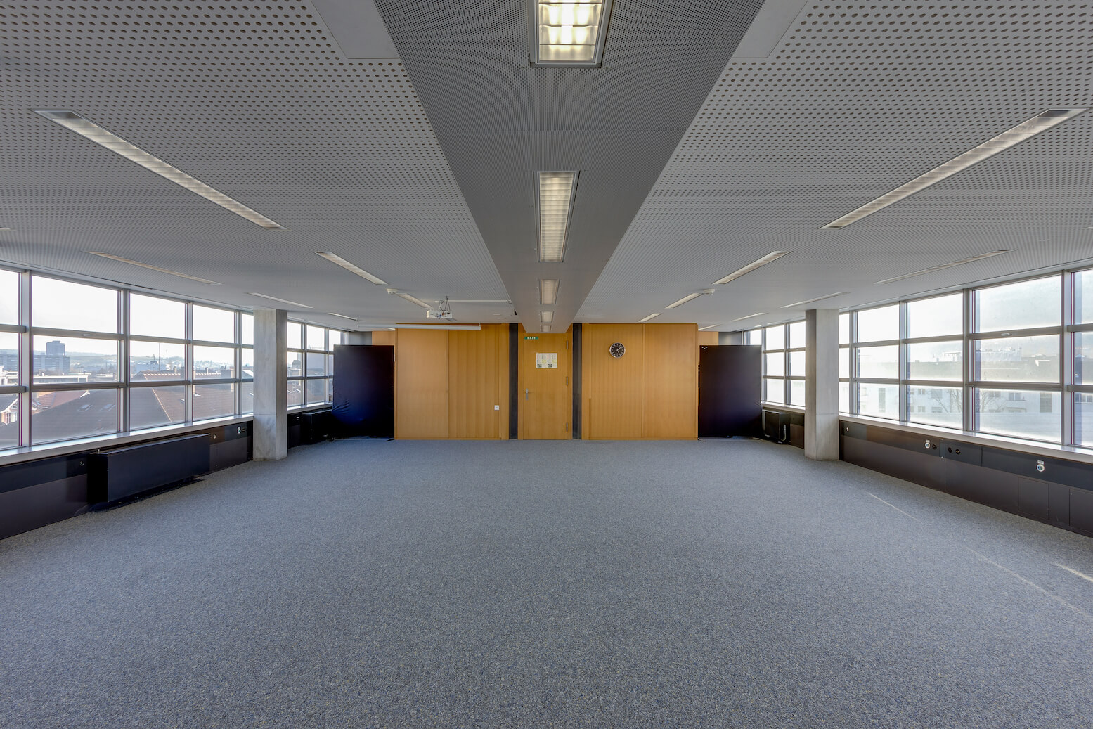 Ein leerer Büroraum mit Teppichboden, großen Fenstern auf drei Seiten und einer holzgetäfelten Wand mit einer Uhr und einer Tür am anderen Ende.
