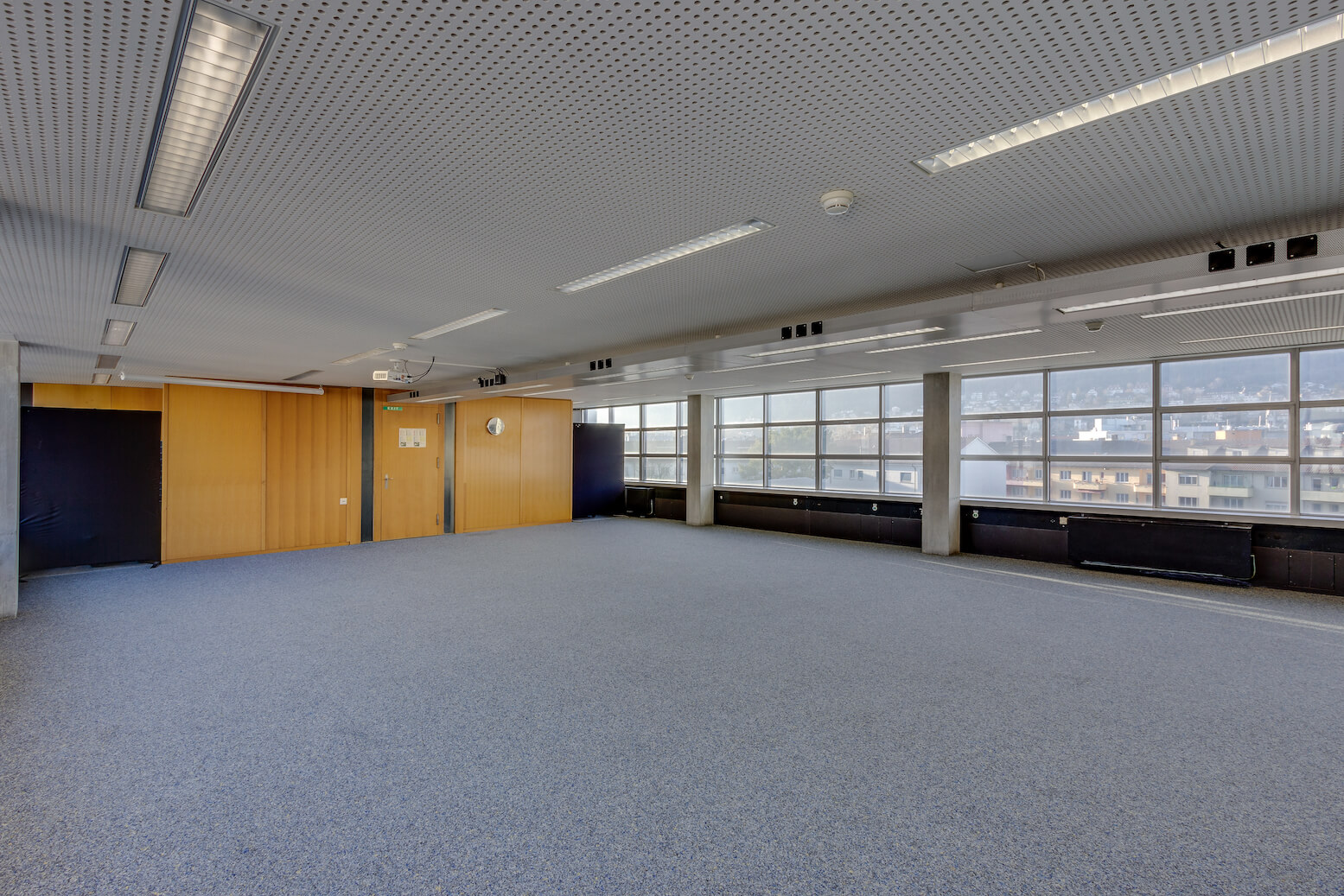 Großer leerer Büroraum mit blauem Teppich, holzvertäfelten Wänden, Deckenleuchten und großen Fenstern mit Blick auf die Stadt.