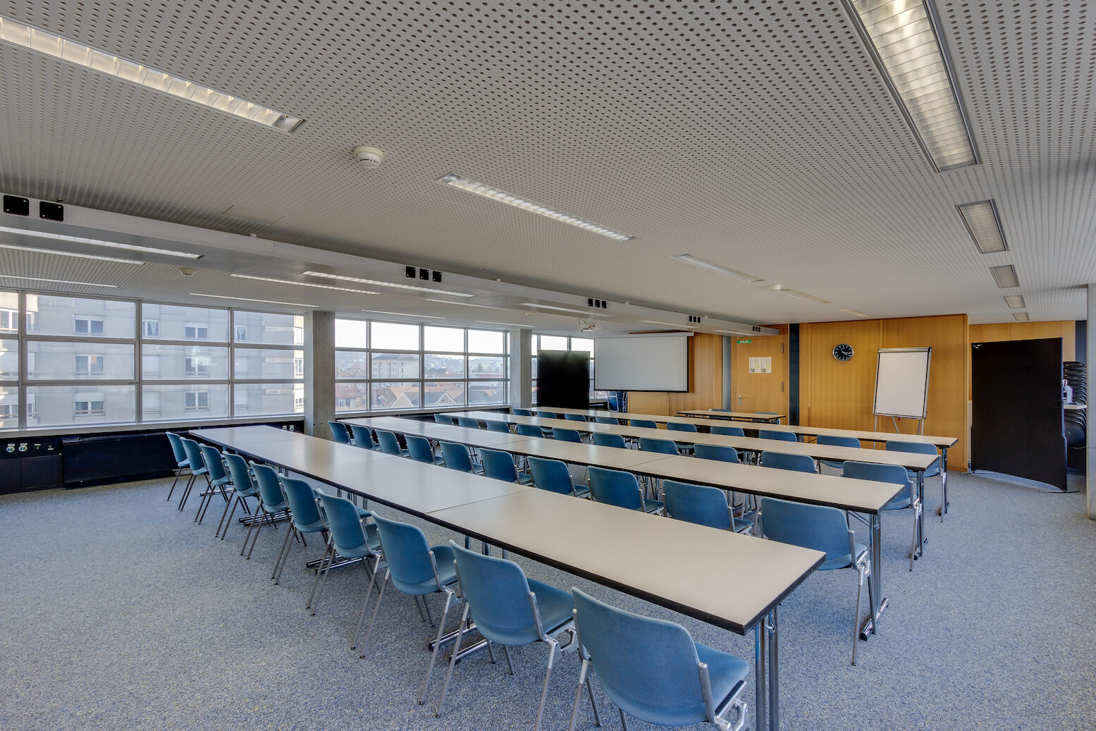 Moderner Konferenzraum mit langen Tischen, blauen Stühlen, großen Fenstern, einem Whiteboard, einer Uhr und einer Projektionsfläche. Der Raum ist leer und gut beleuchtet.