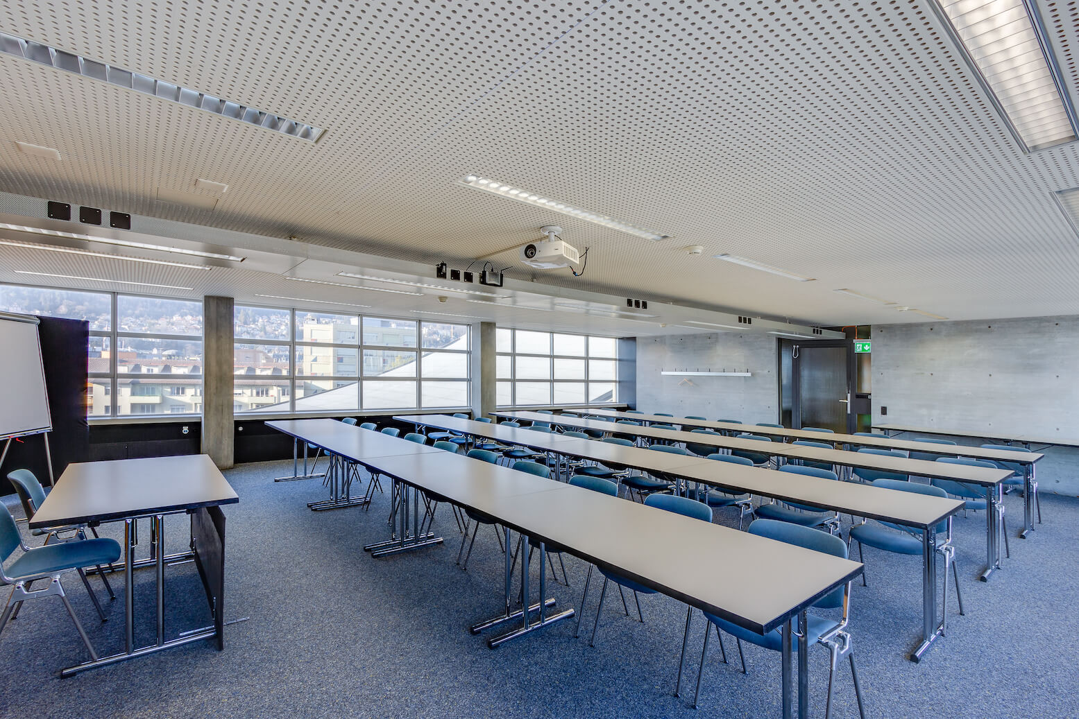 Ein modernes, leeres Klassenzimmer mit Reihen von Tischen und Stühlen vor einer Projektionsfläche, großen Fenstern an einer Wand und heller Deckenbeleuchtung.