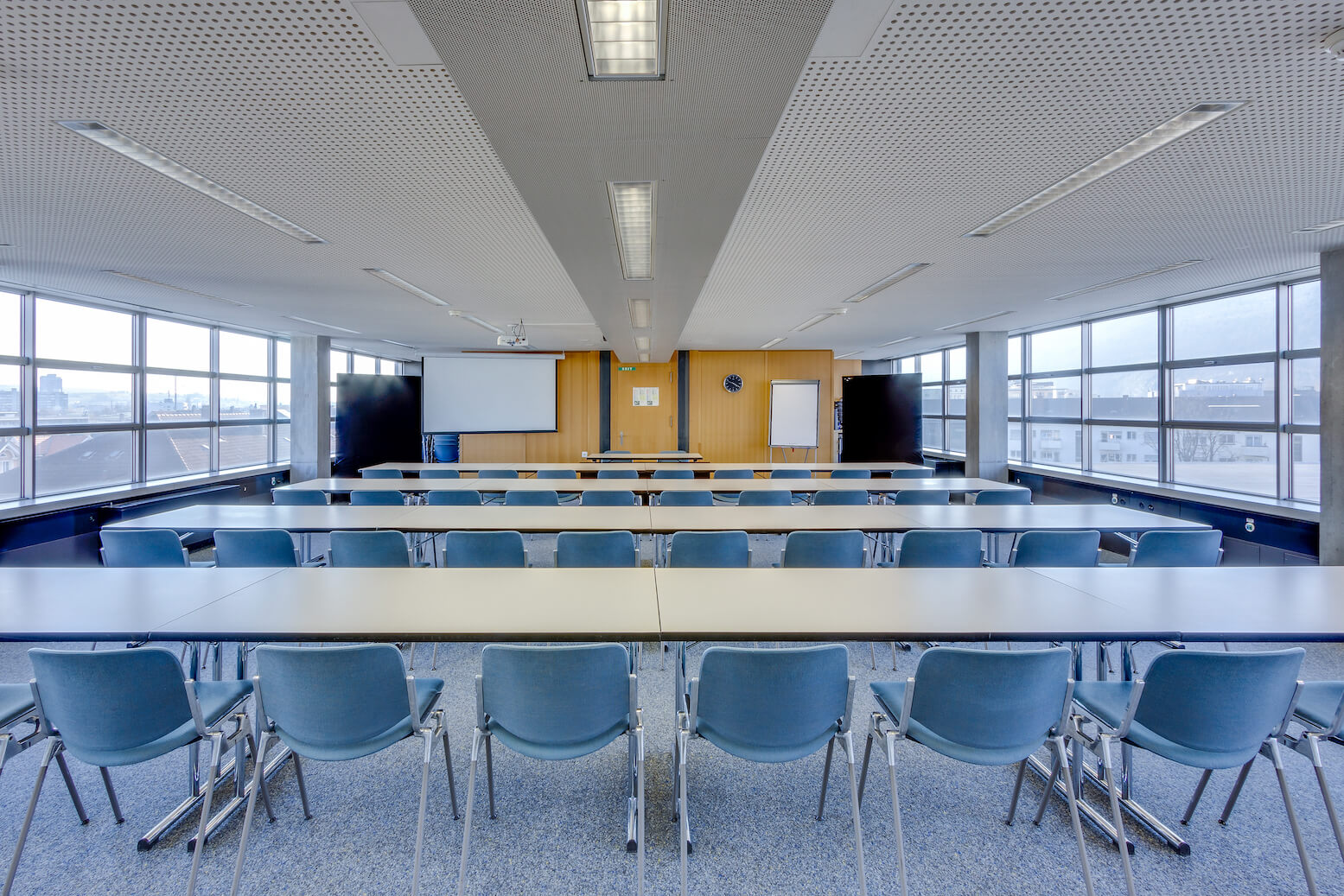Modernes Klassenzimmer mit Reihen blauer Stühle und weißer Tische vor einem Whiteboard, einem Projektor und Fenstern auf beiden Seiten, beleuchtet von Deckenleuchten.