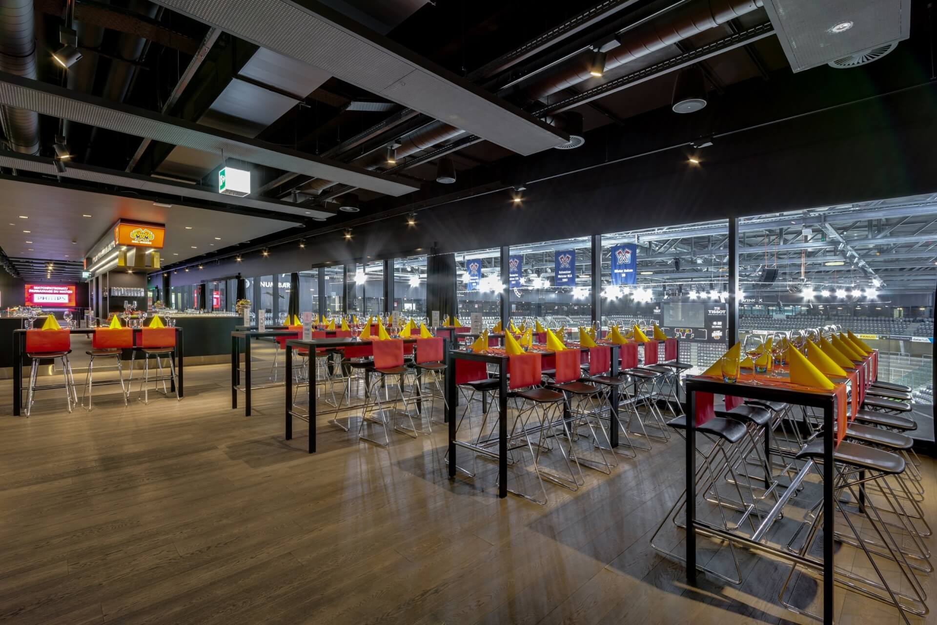 Moderner Essbereich mit Tischreihen, die mit gelben Servietten und roten Behältern gedeckt sind, mit Blick auf eine Sporthalle durch große Glasfenster.