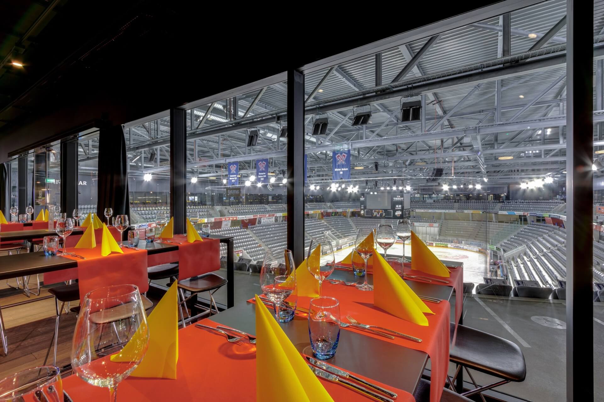 Esstische mit orangefarbenen Tischtüchern und gelben Servietten bieten durch große Glasfenster einen Blick auf eine leere Sporthalle.