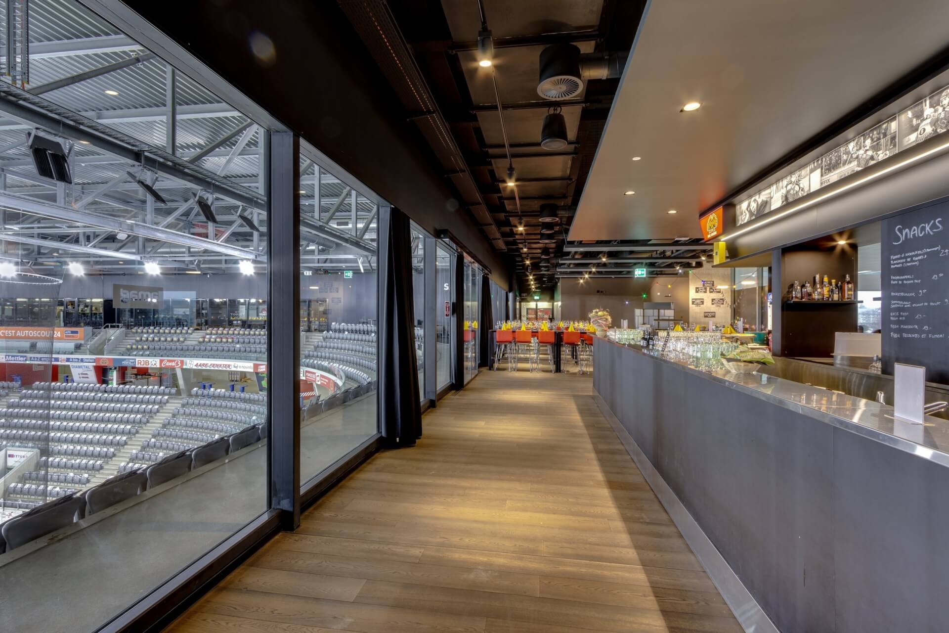 Ein moderner Barbereich mit langem Tresen und Gläsern bietet einen Blick auf eine Sporthalle mit leeren Zuschauerplätzen, die durch große Fenster sichtbar sind.