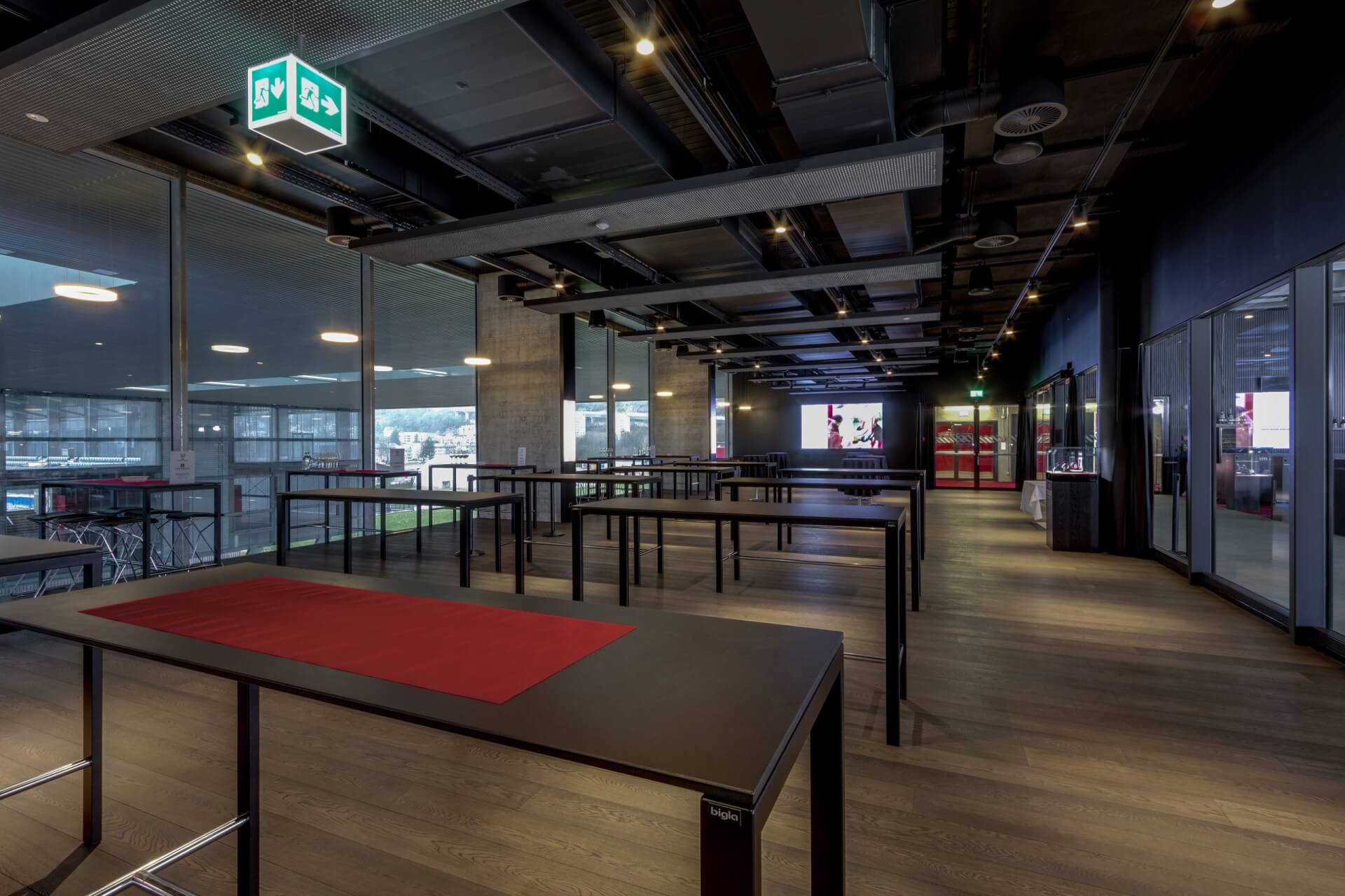 Geräumiger, moderner Raum mit Stehtischen, Holzböden, großen Fenstern und Deckenleuchten, der für Veranstaltungen oder Meetings eingerichtet ist.