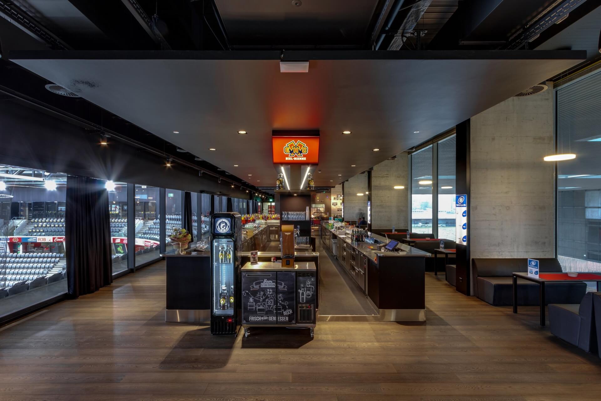 Moderner überdachter Verpflegungsstand mit Lebensmittelauslagen, Kühlschrank, Beschilderung und Sitzgelegenheiten; große Fenster mit Blick auf ein Sportstadion mit leeren Sitzreihen.