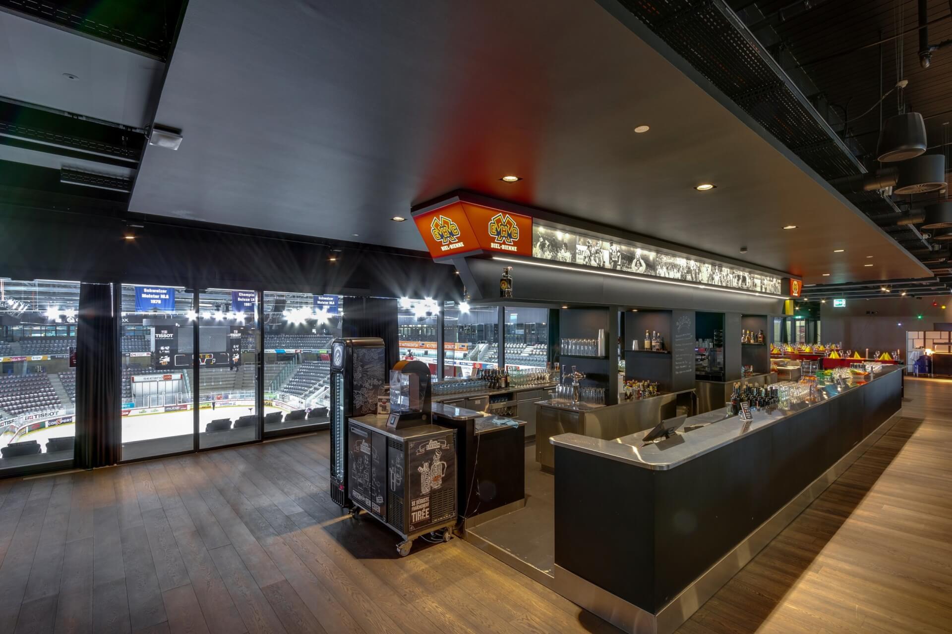 Moderner, überdachter Barbereich mit schlichtem Tresen, ausgestellten Getränken und einem großen Fenster mit Blick auf eine hell erleuchtete Sportarena mit leeren Sitzreihen.