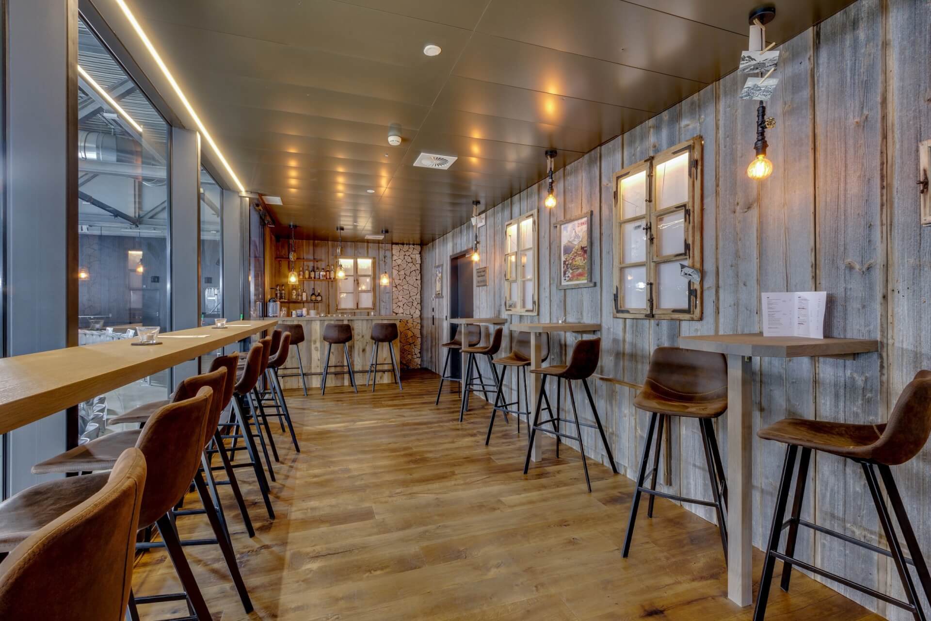 Moderner Barbereich mit Holzböden, hohen Tischen und Stühlen, freiliegenden Glühbirnen, rustikalen holzgetäfelten Wänden und einem kleinen Bartresen im hinteren Teil.