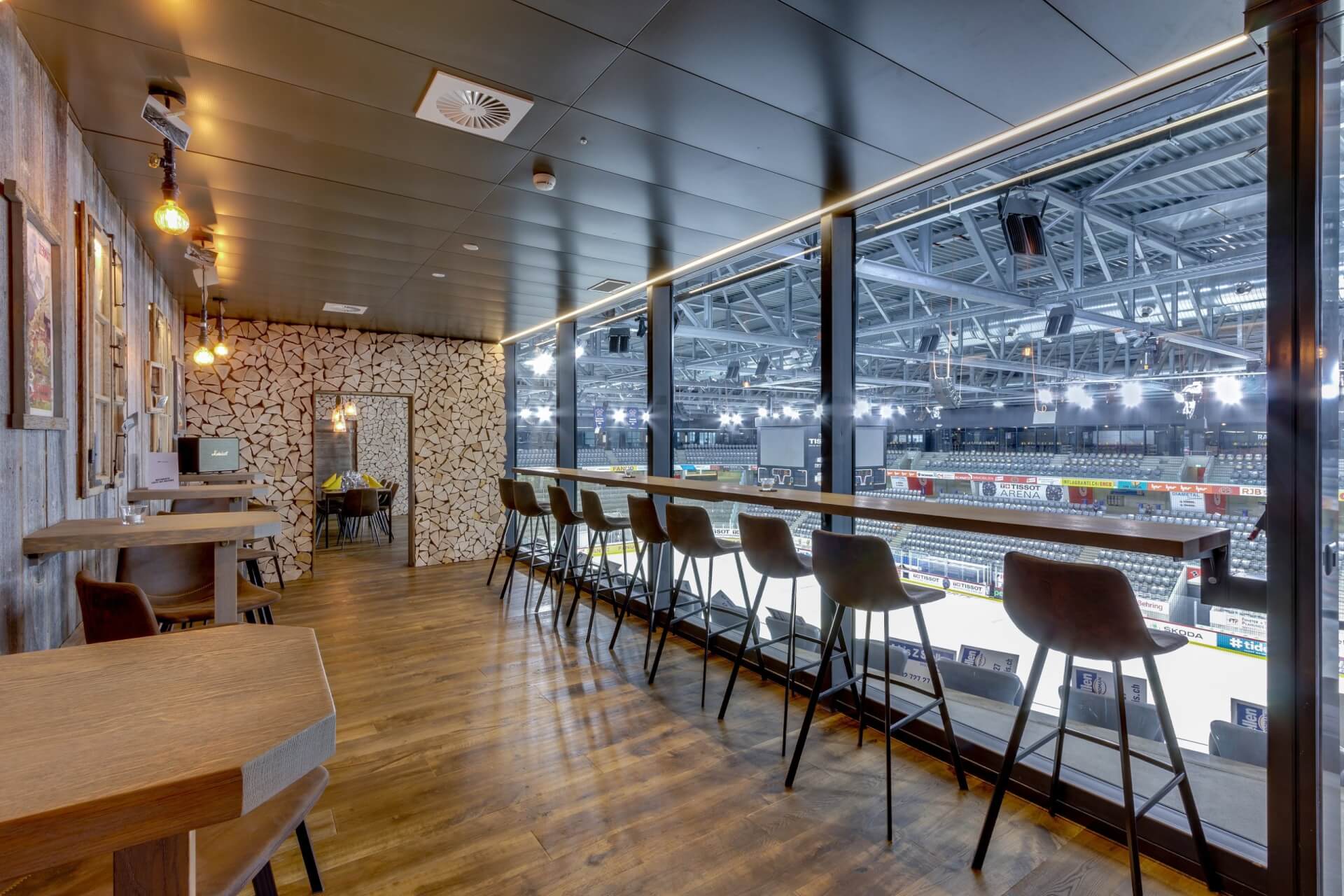 Ein moderner Lounge-Bereich mit Barhockern und Tischen bietet durch große Glasfenster einen Blick auf eine Sporthalle. Der Raum ist mit Holzböden und modernem Dekor ausgestattet.