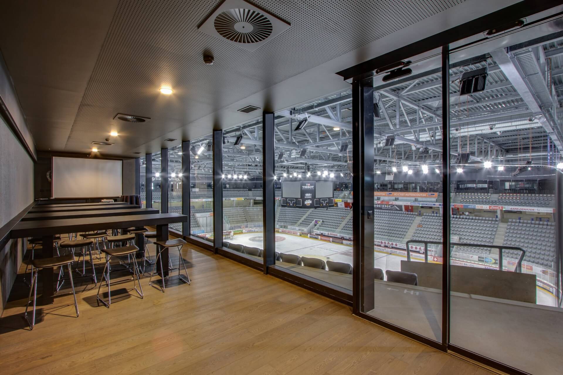 Eine moderne Suite mit Glaswänden bietet einen Blick auf eine leere Eishockeyhalle, mit Sitzreihen und der darunter liegenden Eisfläche.