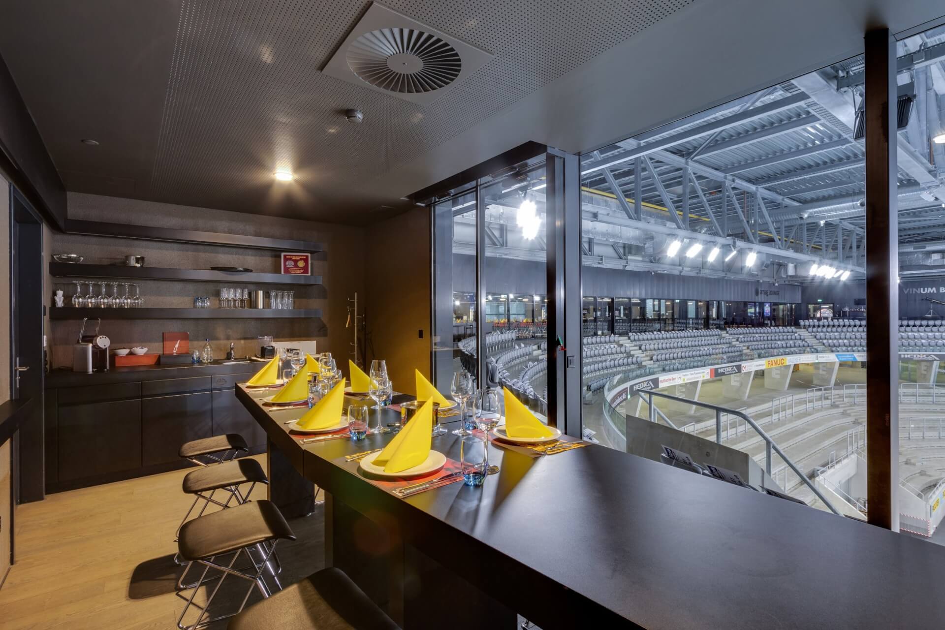 Ein privater Essbereich mit einem gedeckten Tisch und gelben Servietten bietet einen Blick auf eine leere Sporthalle mit abgestuften Sitzgelegenheiten und heller Deckenbeleuchtung.