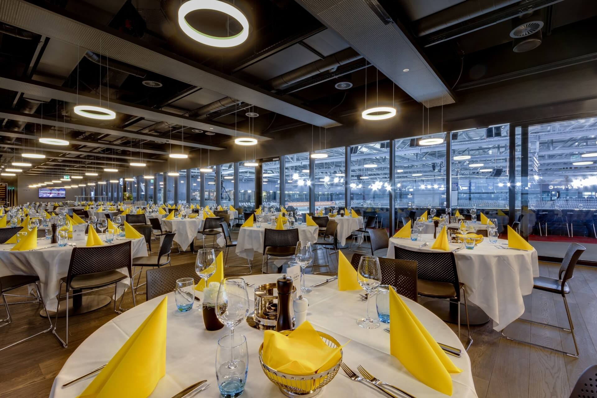 Ein moderner Essbereich mit runden Tischen, die mit weißen Tischdecken, gelben Servietten, Gläsern und Besteck eingedeckt sind, mit Blick durch Glasfenster auf eine große Sporthalle.