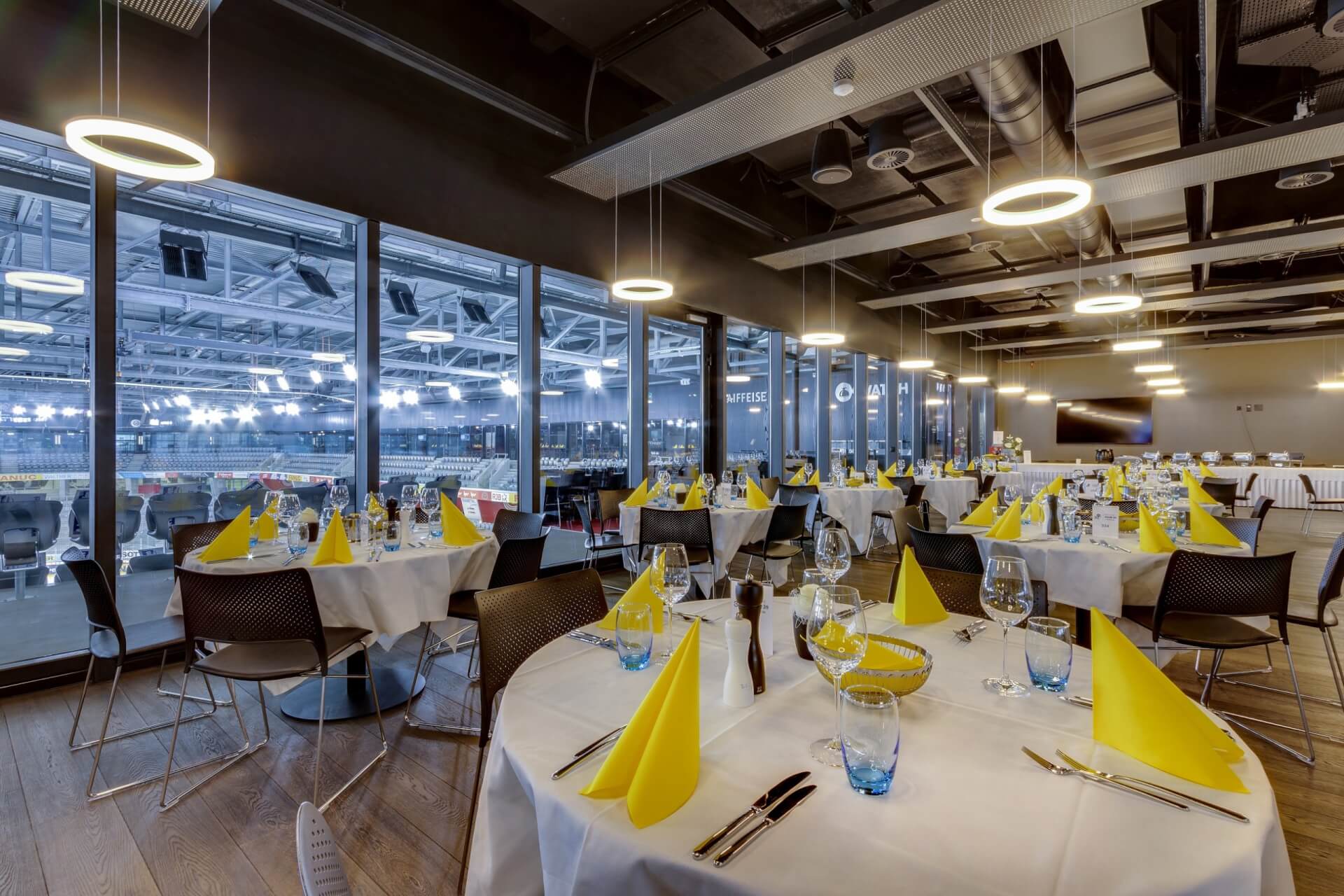 Moderner Essbereich mit runden Tischen, die mit gelben Servietten und Gläsern gedeckt sind, mit Blick auf ein Sportstadion durch große Fenster.