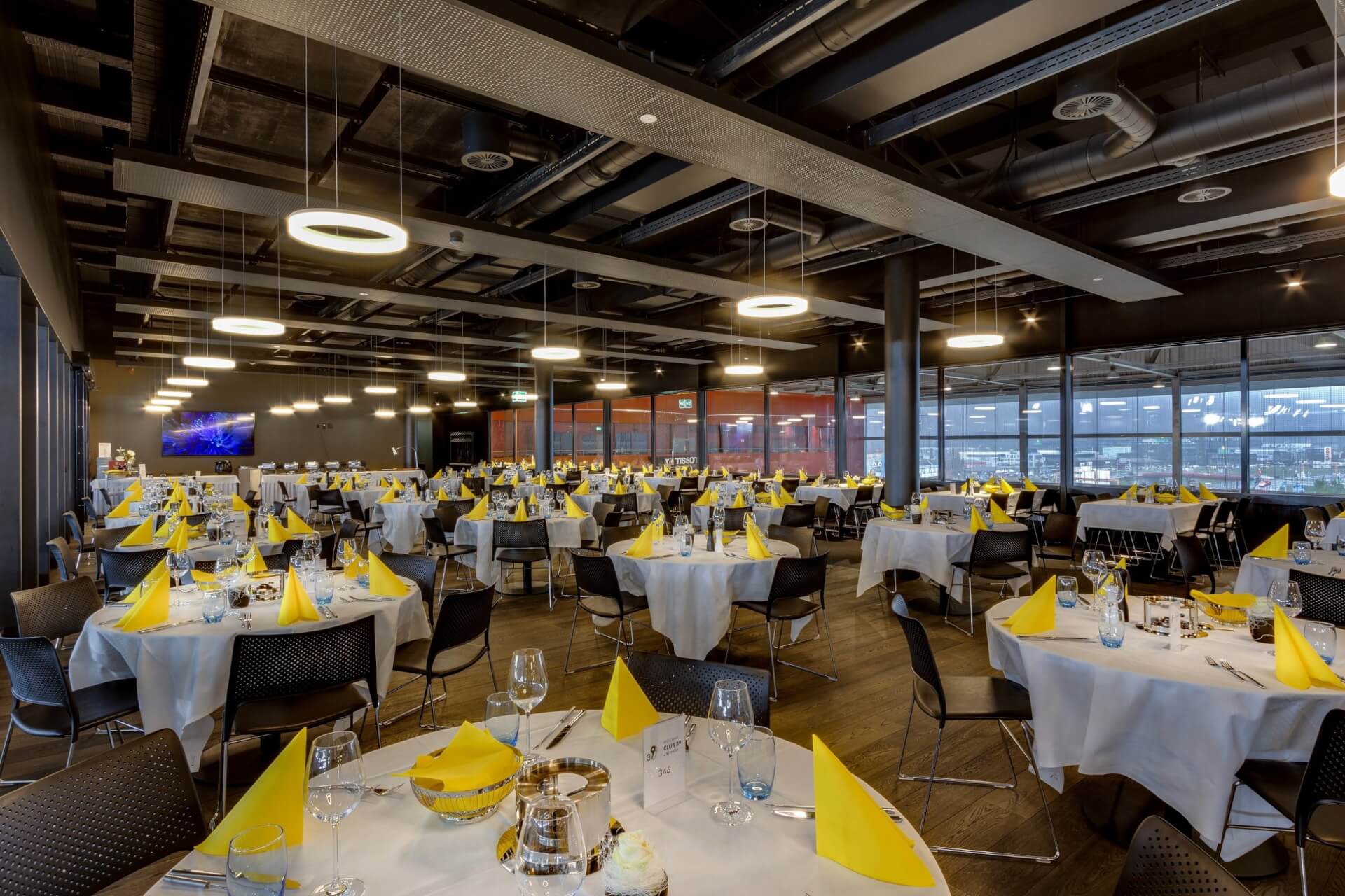 Geräumiger, moderner Speisesaal mit runden Tischen mit weißen Tüchern und gelben Servietten, schwarzen Stühlen und großen Fenstern an einer Wand. An der Decke befinden sich freiliegende Rohre und runde Leuchten.
