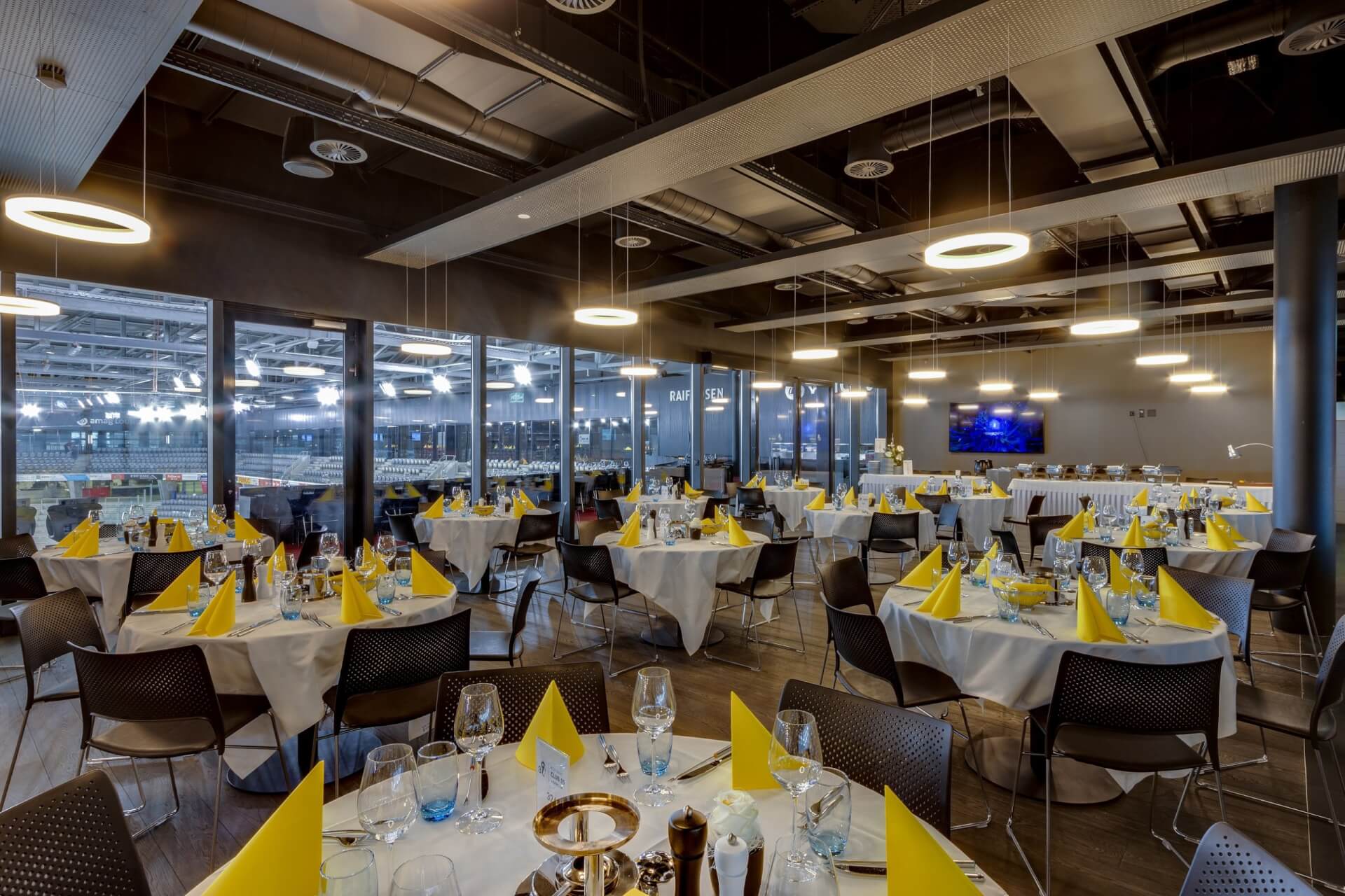 Ein moderner Speisesaal mit runden Tischen, die für eine Veranstaltung gedeckt sind, mit gelben Servietten, Gläsern und kreisförmigen Deckenleuchten. Große Fenster bieten einen Blick auf eine Sporthalle.